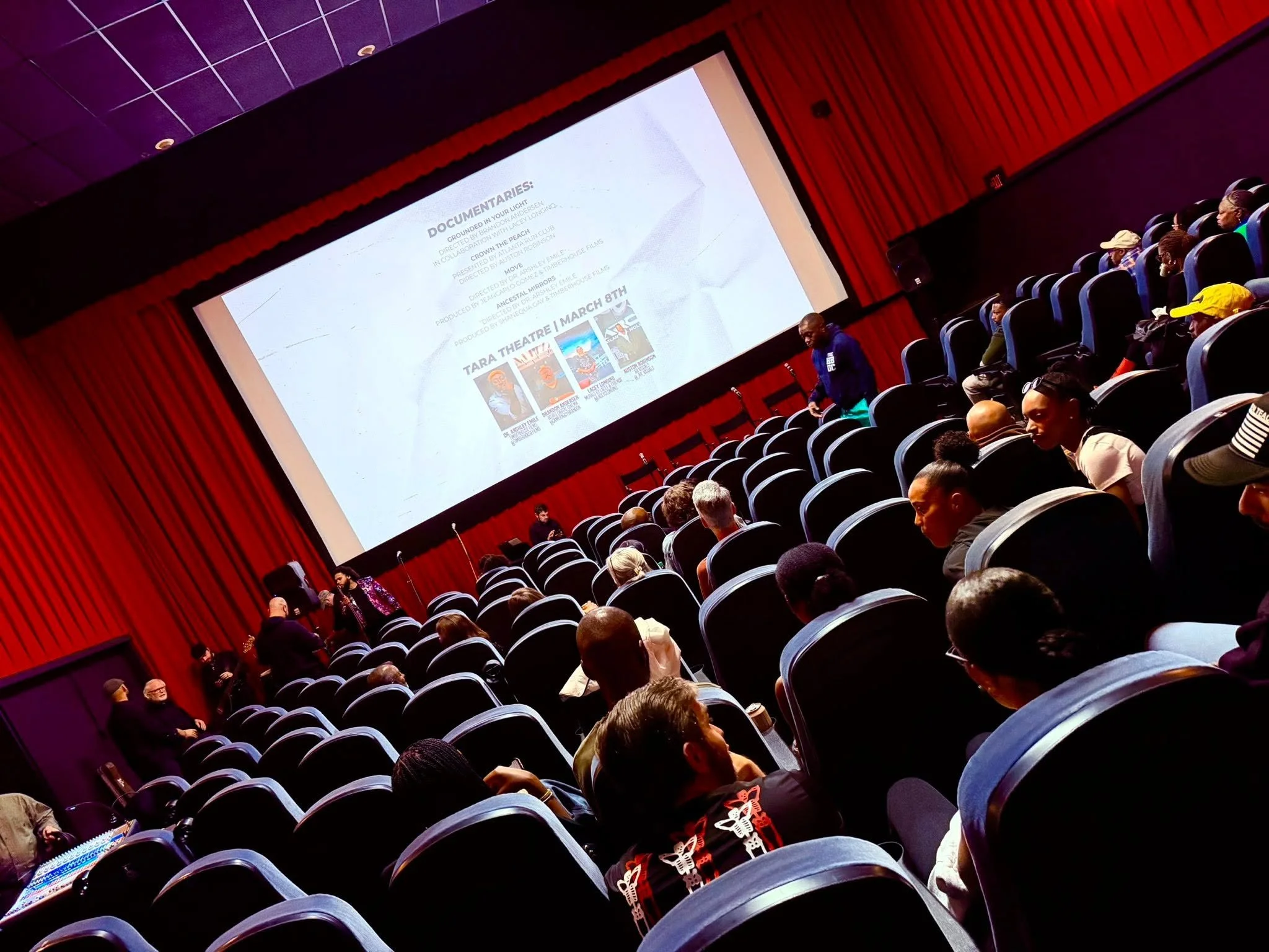 Audience seated in theater seats watching a presentation on a large screen, with some people walking and chatting.
