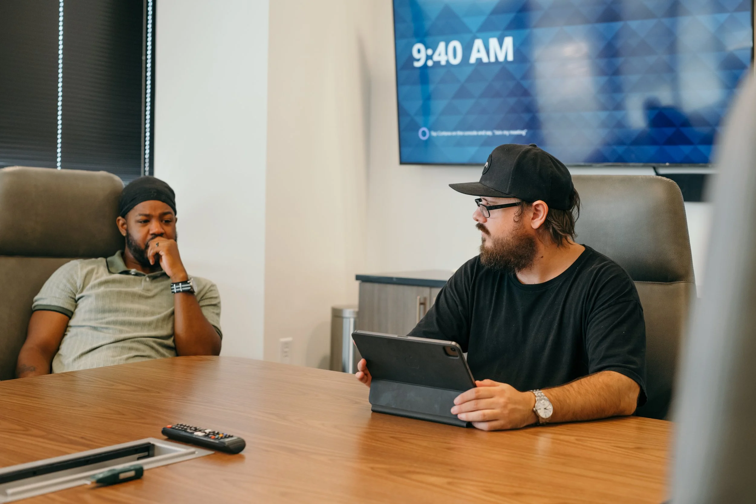 Two men sit at a conference table in a modern office, engaged in a discussion. One man, wearing a light-colored polo shirt and a black headband, appears to be listening thoughtfully. The other man, with glasses, a black cap, and a beard, is holding a tablet and looking at the man in the polo shirt. On the table are remote controls and a closed laptop. In the background, a large screen displays the time, 9:40 AM.
