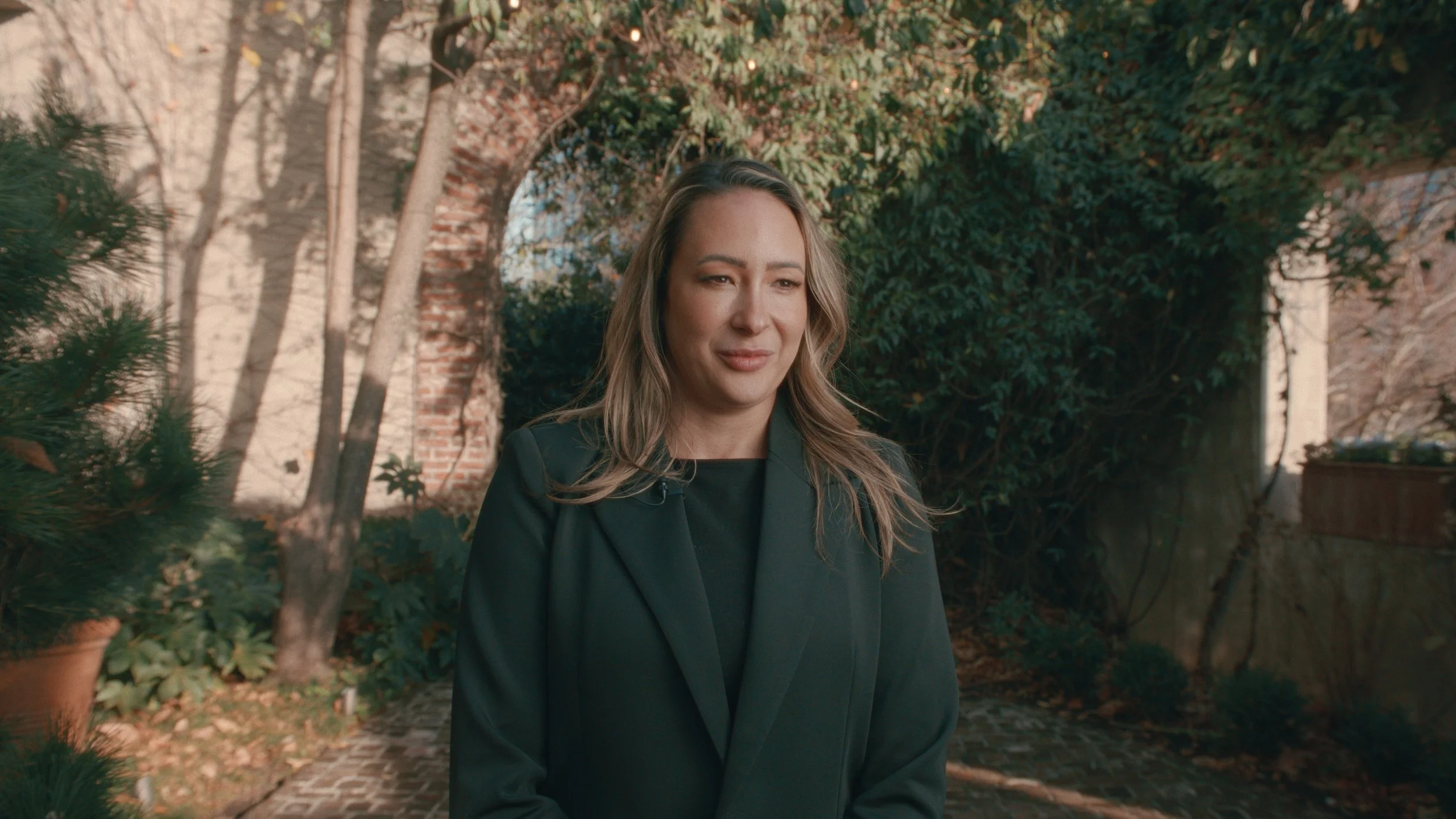 A woman with wavy blonde hair wearing a black blazer. She is standing outdoors in front of a brick wall, trees, and shrubbery on a sunny day.