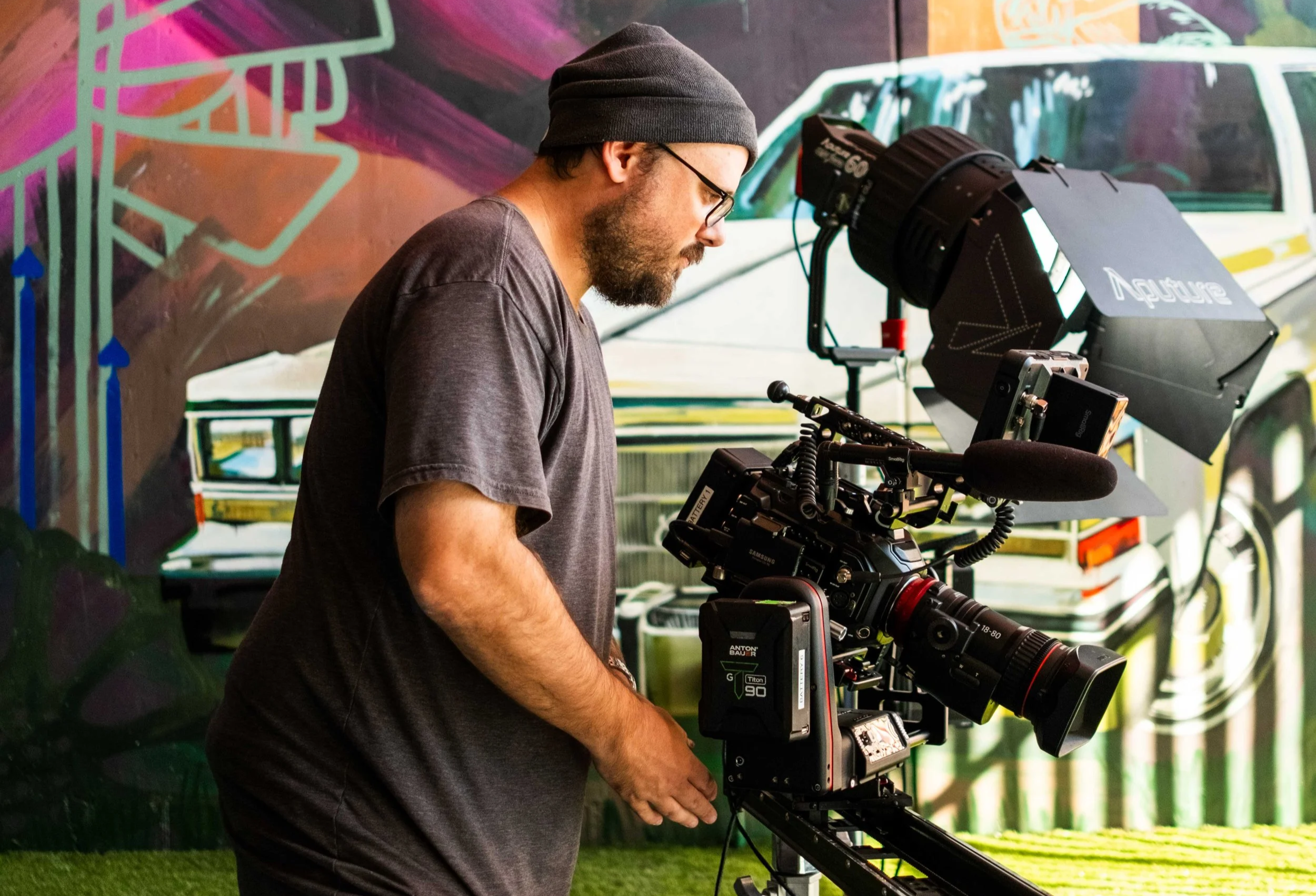 A man wearing glasses and a beanie operating a professional camera on a tripod, with a colorful mural in the background.