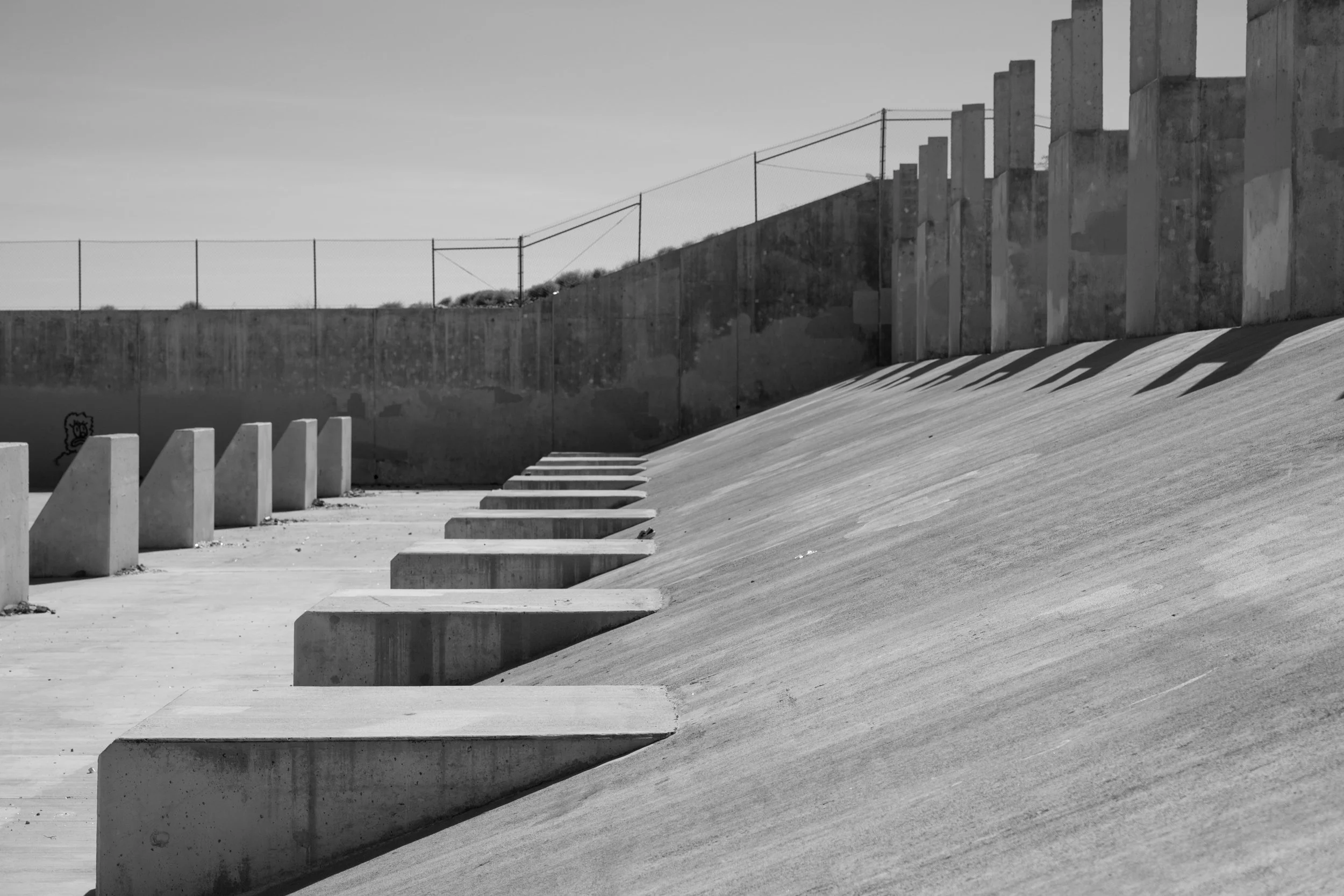 Black and white photo of a concrete staircase and a retaining wall, with a chain-link fence on top. Shadows cast on the ground and wall.