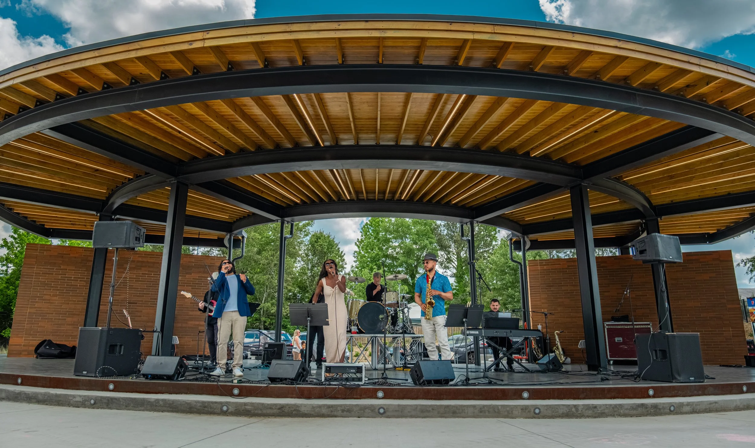 Musicians performing on an outdoor stage with a wooden roof, trees, and a cloudy sky in the background.