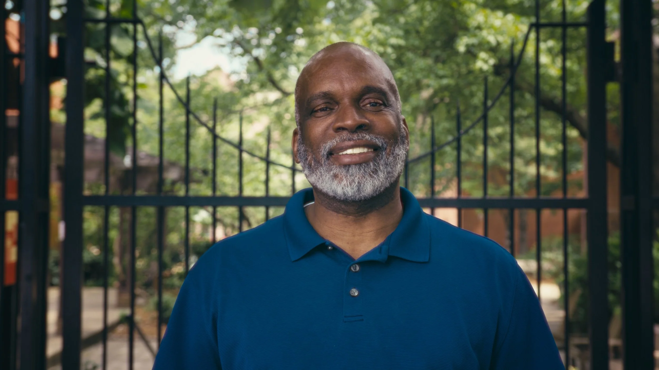 Close-up of a happy middle-aged man with a gray beard, smiling, wearing a blue polo shirt, standing in front of a black metal fence with green trees in the background.