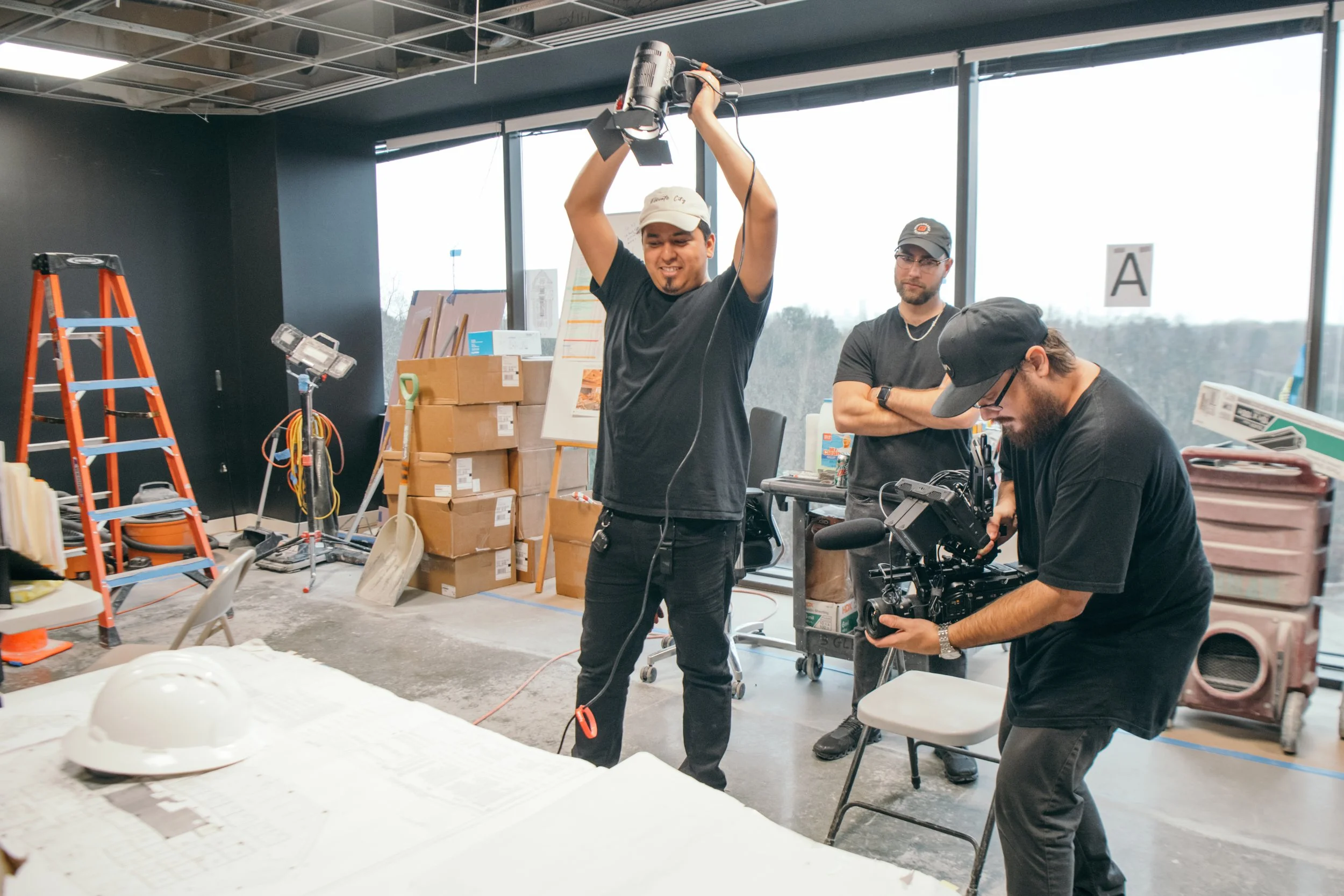 People working in a studio or workshop, one man holding lighting equipment, another man looking at a camera, with boxes, tools, and equipment around.