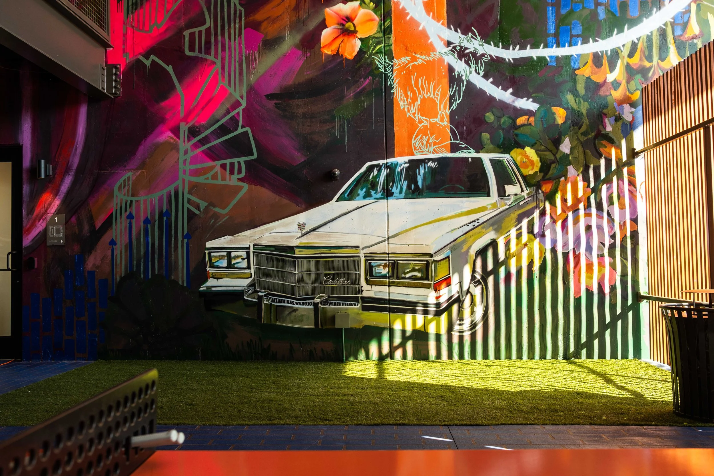Colorful mural on a wall featuring a white vintage Cadillac, orange flowers, vibrant abstract patterns, and green foliage.