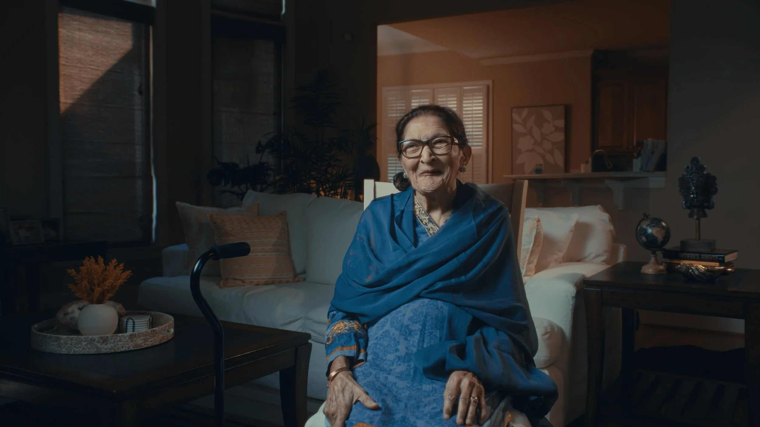 An elderly woman with glasses smiling and sitting in a living room with sunlight coming through the window, decorated with plants and artwork
