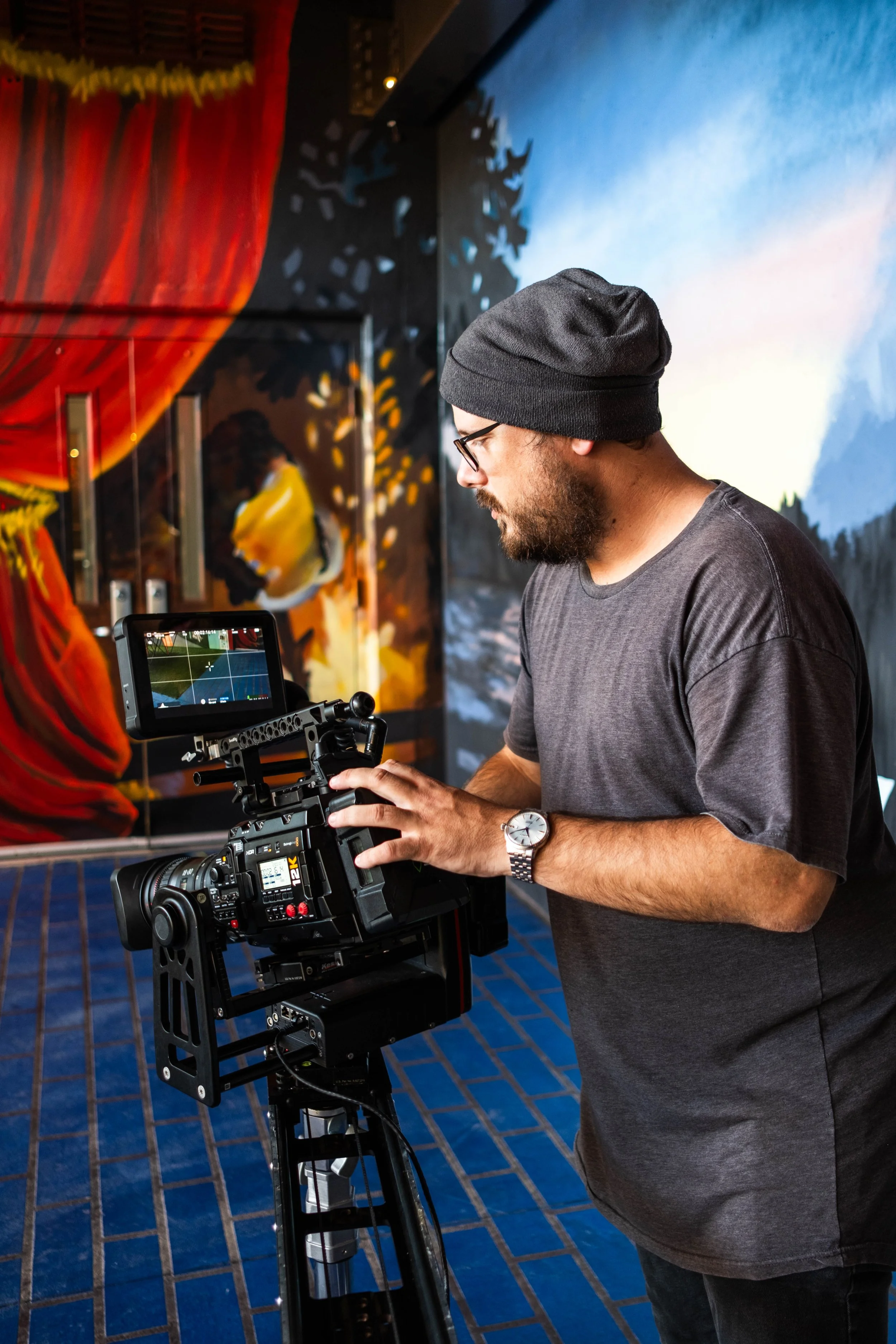 A man with glasses, a gray T-shirt, a black beanie, and a watch is operating a professional video camera on a tripod in a room with colorful murals.