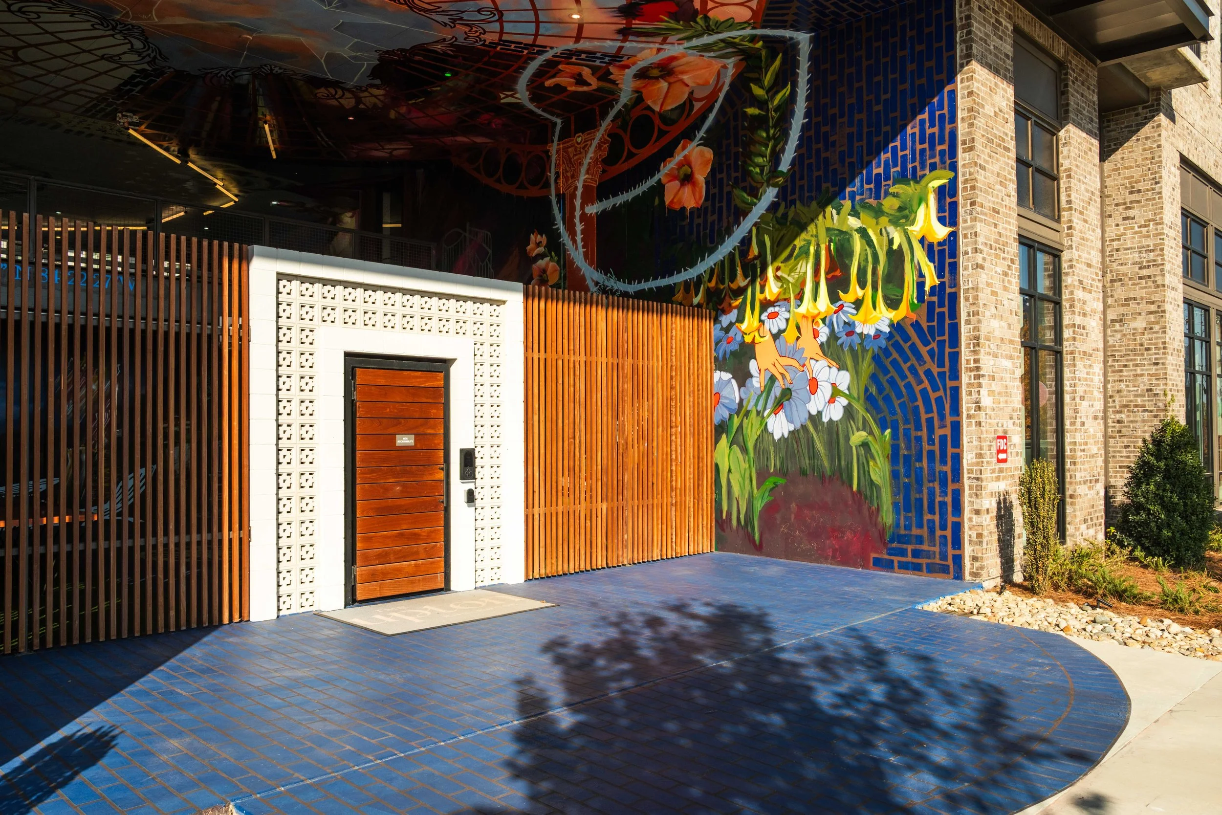 Colorful mural on building wall with flowers and barbed wire design, brick and wood exterior, decorative fence, tiled sidewalk, and nearby greenery.