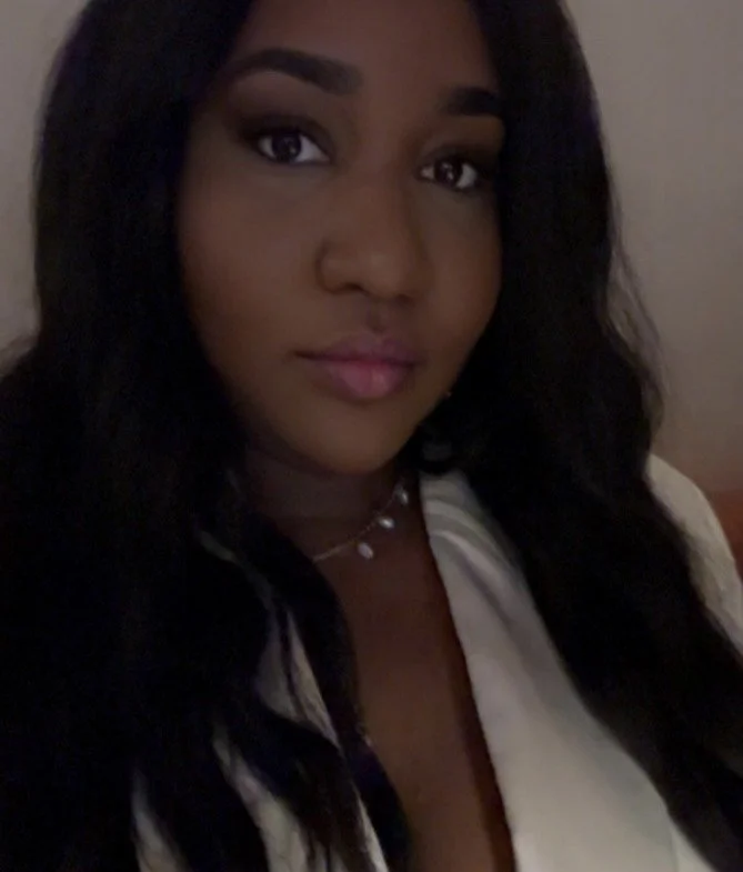 Close-up of a young woman with long dark hair, wearing a white top and a delicate necklace, looking at the camera with a slight smile.