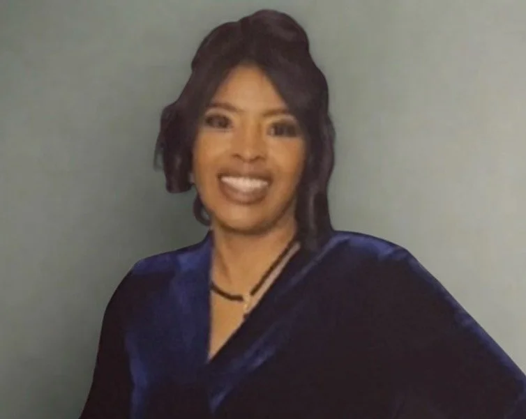 A woman with dark hair smiling, dressed in a dark blue satin blouse and layered necklaces, standing against a plain light-colored background.