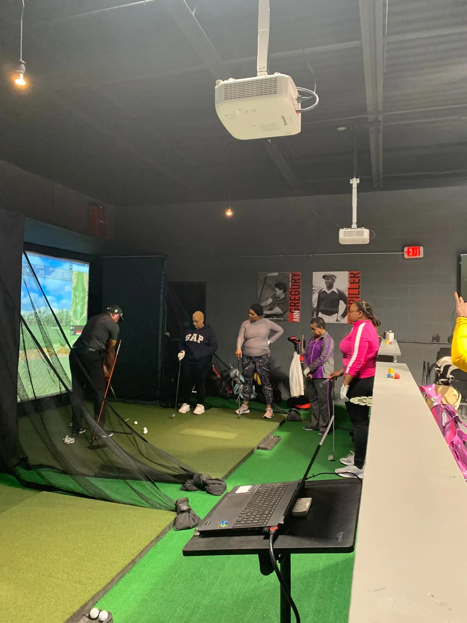 Indoor golf practice area with people practicing and watching, with a golf course simulator screen, golf clubs, and equipment.