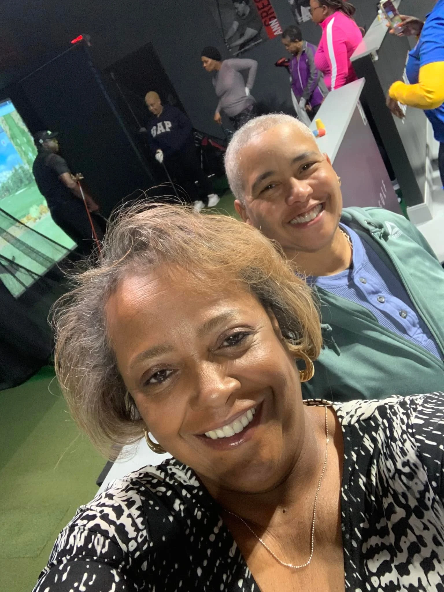 Two women taking a selfie at a mini golf course, with other people in the background playing mini golf and using their phones.