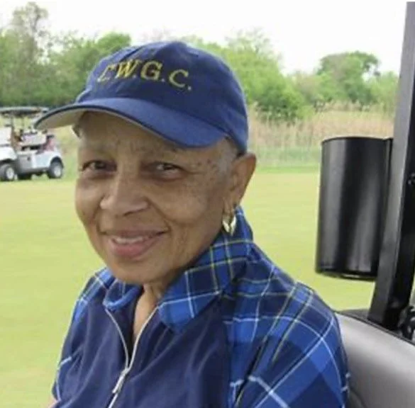 An elderly woman smiling, wearing a blue plaid shirt and a navy cap with yellow letters. She is sitting on a golf cart on a golf course with trees and a golf cart in the background.