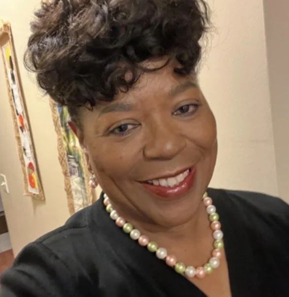 A woman smiling, wearing a black top and a colorful pearl necklace, with short curly hair and earrings.