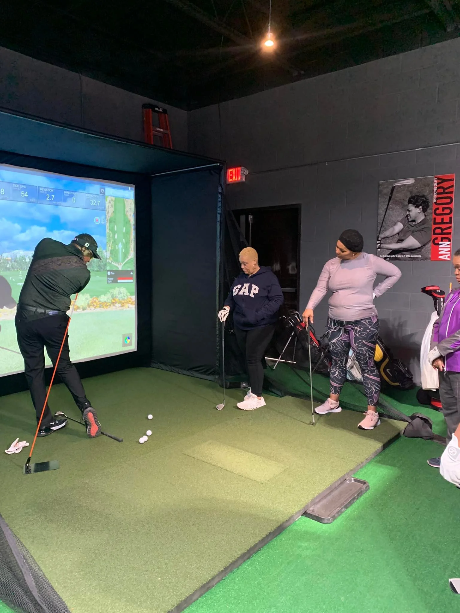 Three women and a man practicing golf on an indoor simulator, with the man preparing to hit the golf balls.