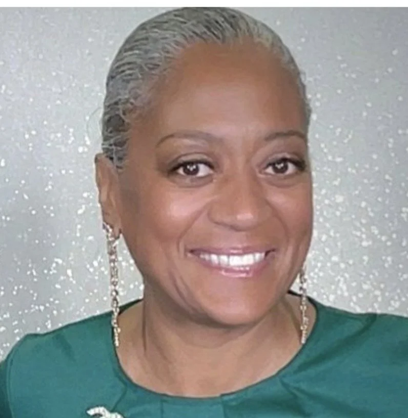 A smiling woman with short gray hair, wearing earrings and a green top, standing in front of a sparkly silver background.