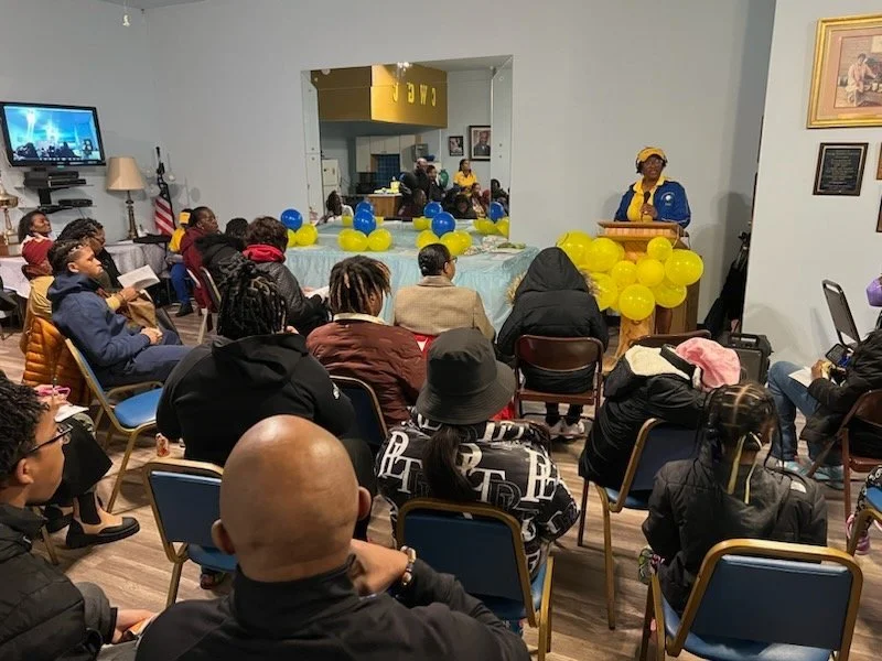 A woman wearing a blue vest and yellow hat delivers a speech at a podium decorated with yellow balloons, with an audience of diverse adults and children listening attentively in a community room decorated with blue and yellow balloons.