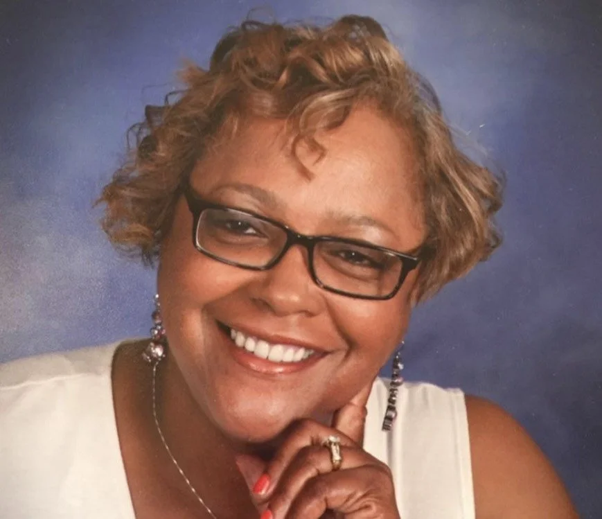 Close-up portrait of a joyful woman with curly light brown hair, glasses, and earrings, smiling with her hand resting on her chin against a blue background.
