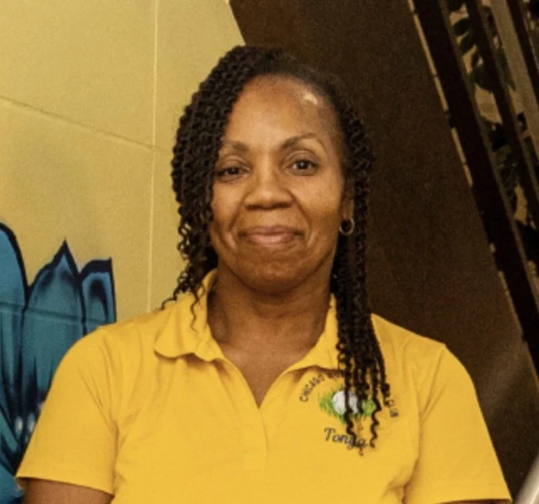 A woman with braided hair wearing a yellow polo shirt standing indoors with graffiti art in the background.