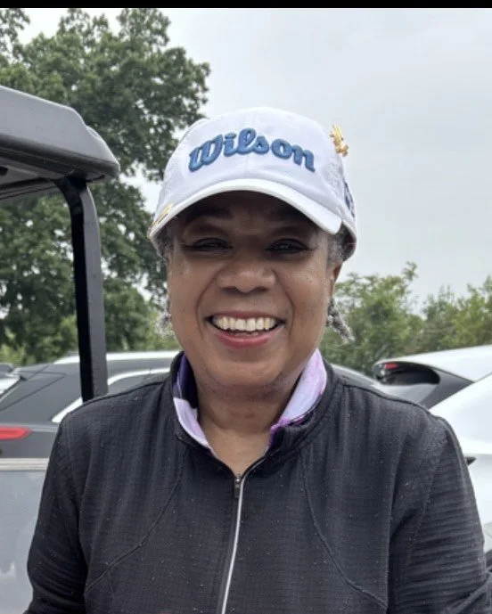 A smiling woman wearing a white Wilson cap and black jacket outdoors with cars and trees in the background.