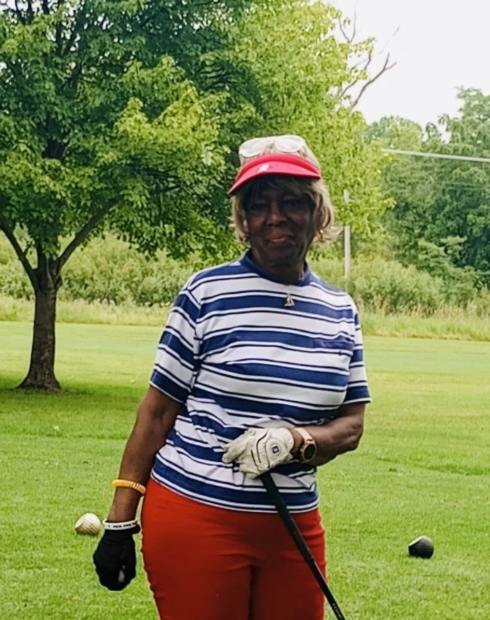 An elderly woman on a golf course holding a golf club, wearing a red visor, striped blue and white shirt, red pants, and a white golf glove, with trees and green grass in the background.