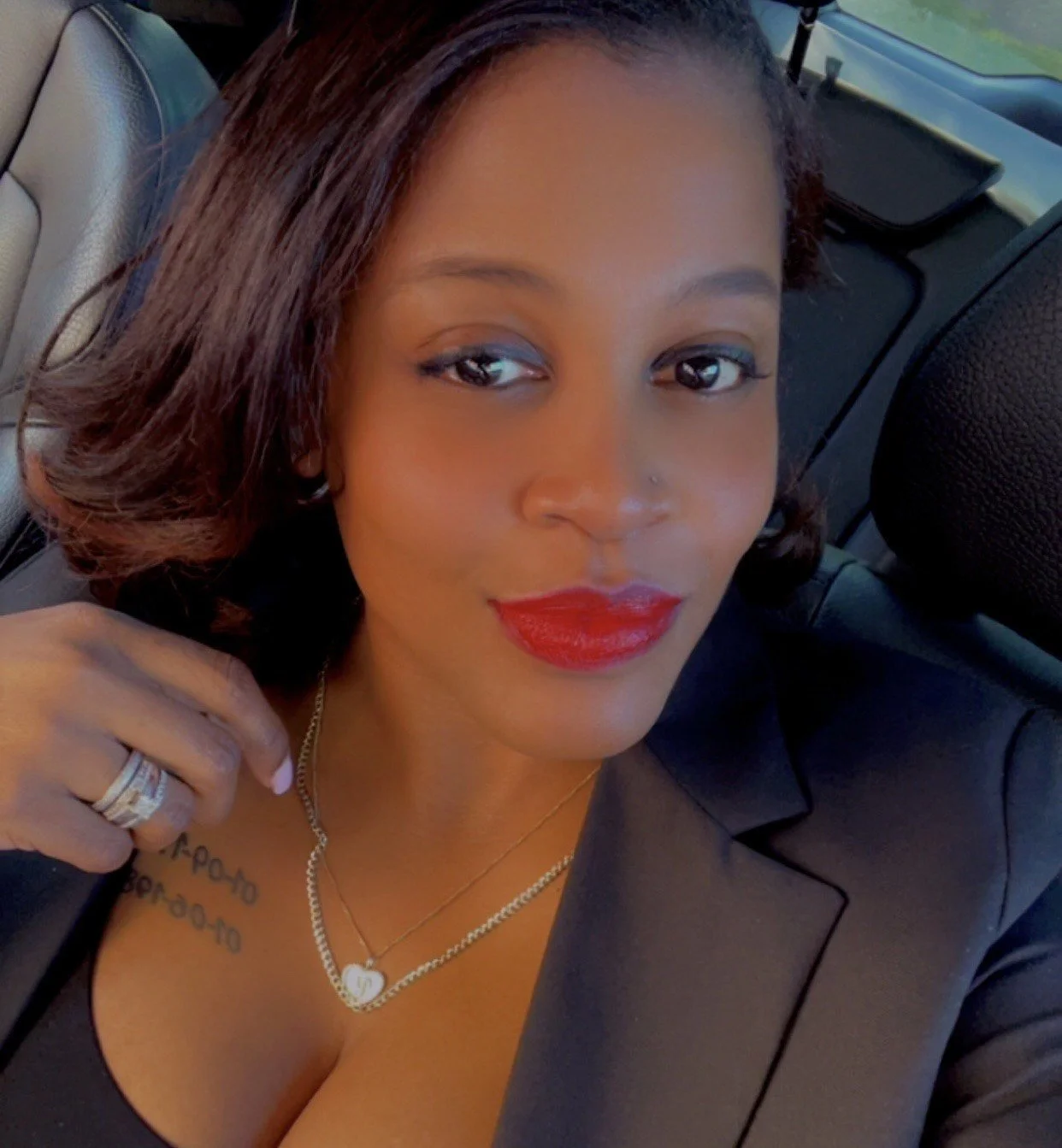 A woman with shoulder-length dark hair, red lipstick, wearing a black outfit and jewelry, sitting inside a car.