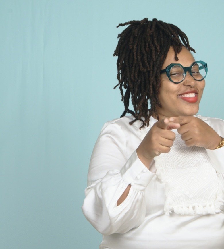 A woman with glasses, dreadlocks, and red lipstick pointing and smiling at the camera.