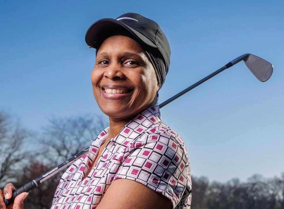 Smiling woman wearing a black cap and patterned golf shirt holding a golf club on a golf course.