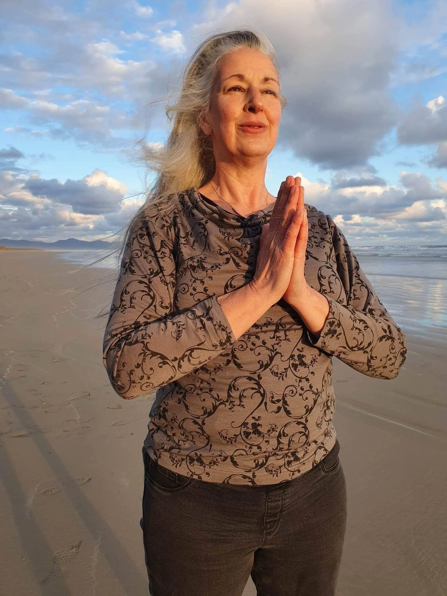 A woman practicing yoga on the beach during sunset, with her hands pressed together in a prayer position and her eyes gently closed.