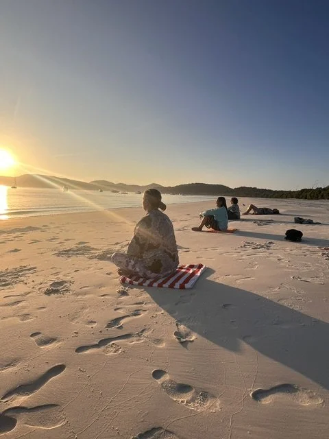 People relaxing on a sandy beach at sunset, sitting on towels, with footprints in the sand, overlooking the ocean and distant islands.