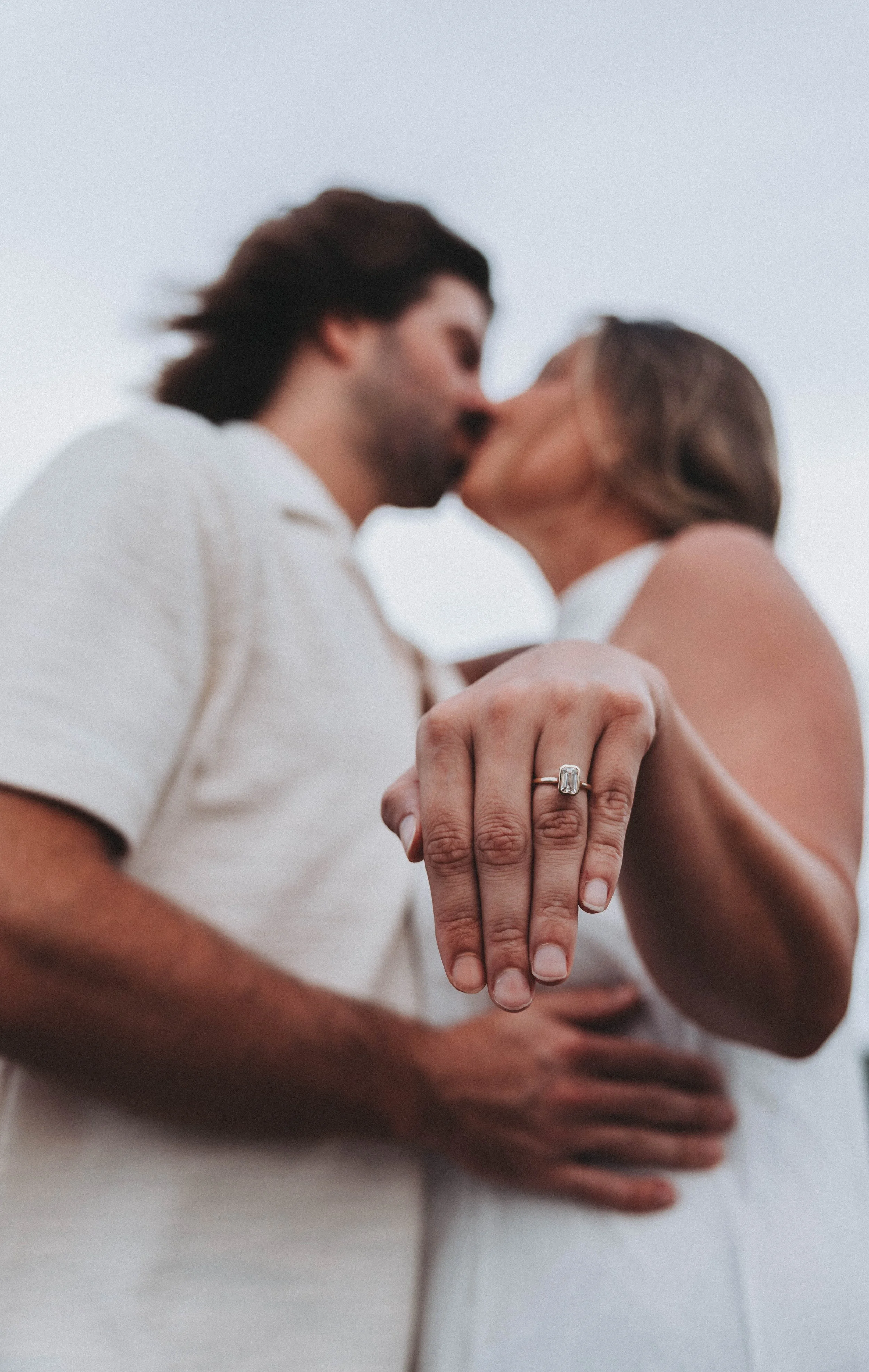 Close-up of a couple kissing with a focus on a woman's hand wearing an engagement ring, against a clear sky background.