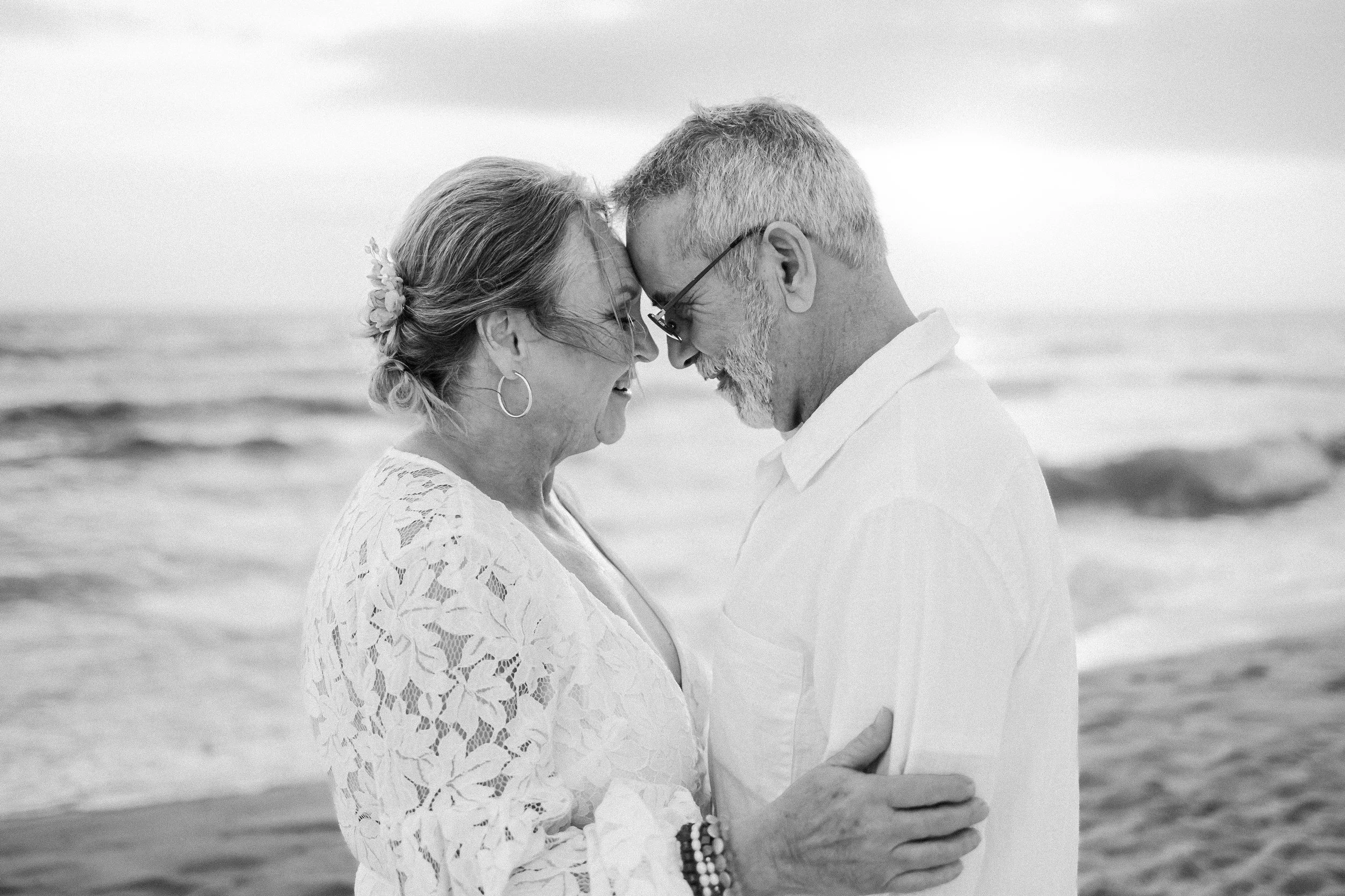 Elderly couple embracing on a beach, forehead to forehead, smiling, with ocean in the background.