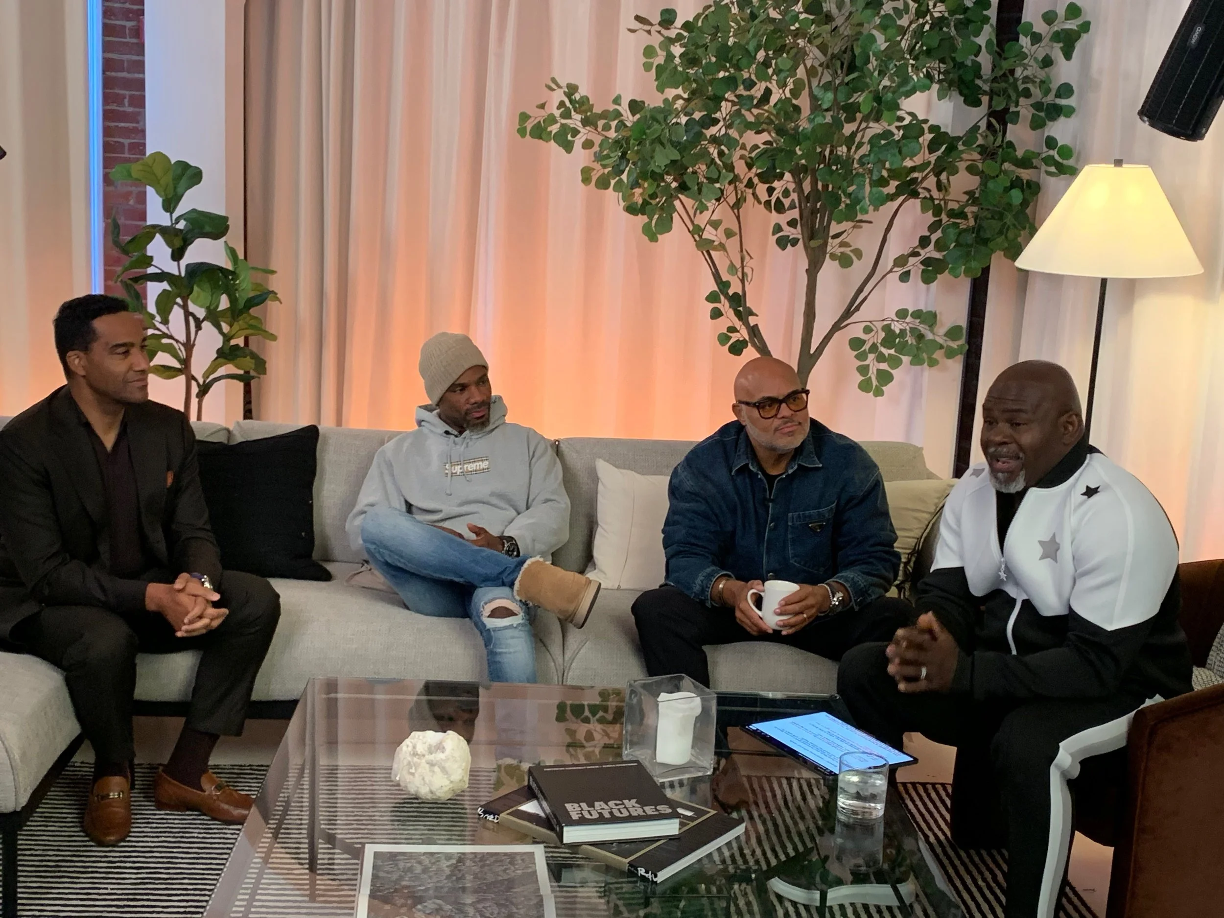 Four men sitting on a beige couch and a chair in a living room, engaged in conversation. They are in front of a glass coffee table with books, a candle, and a stone sculpture. Behind them are curtains, large potted plants, and a standing lamp.