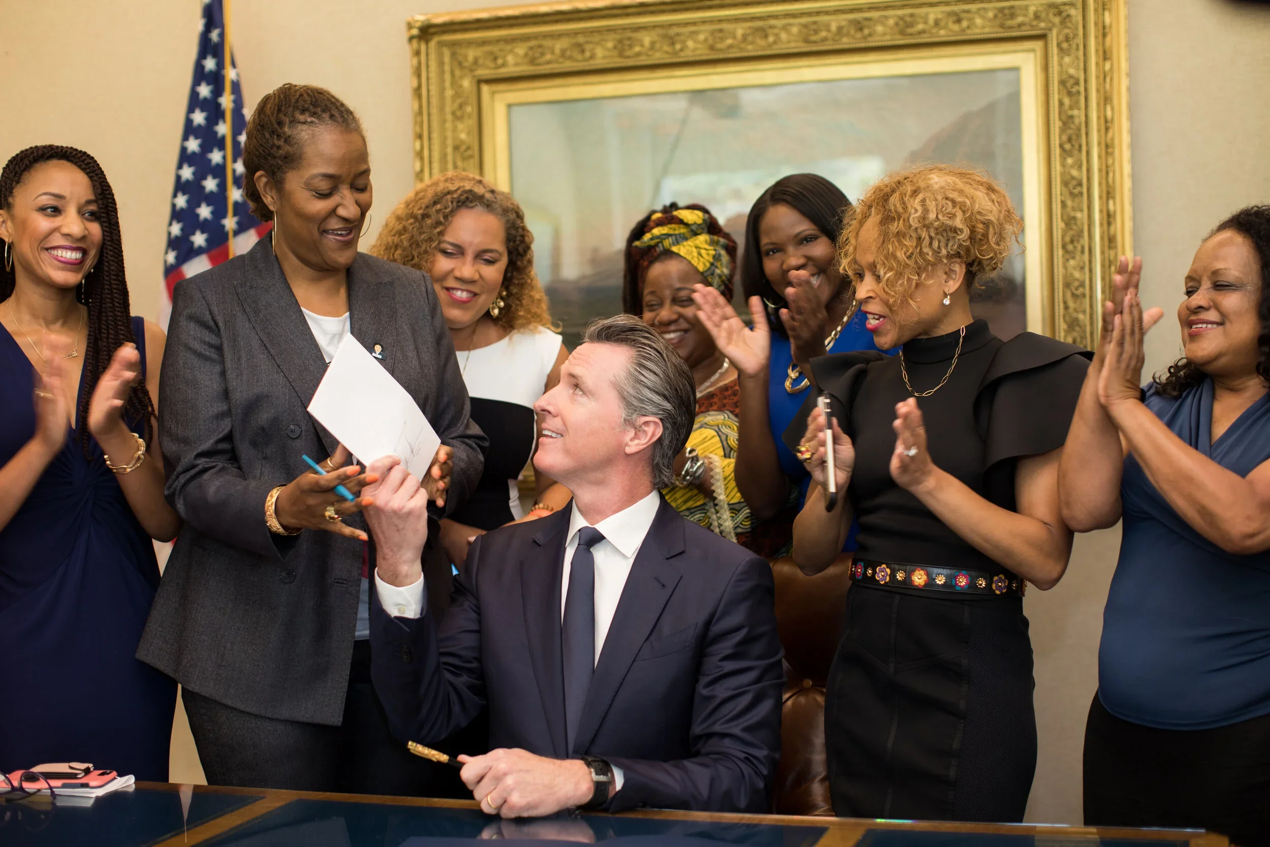 A man in a suit sitting at a desk, surrounded by women cheering and smiling, as the woman in a gray blazer signs a document, with an American flag in the background.
