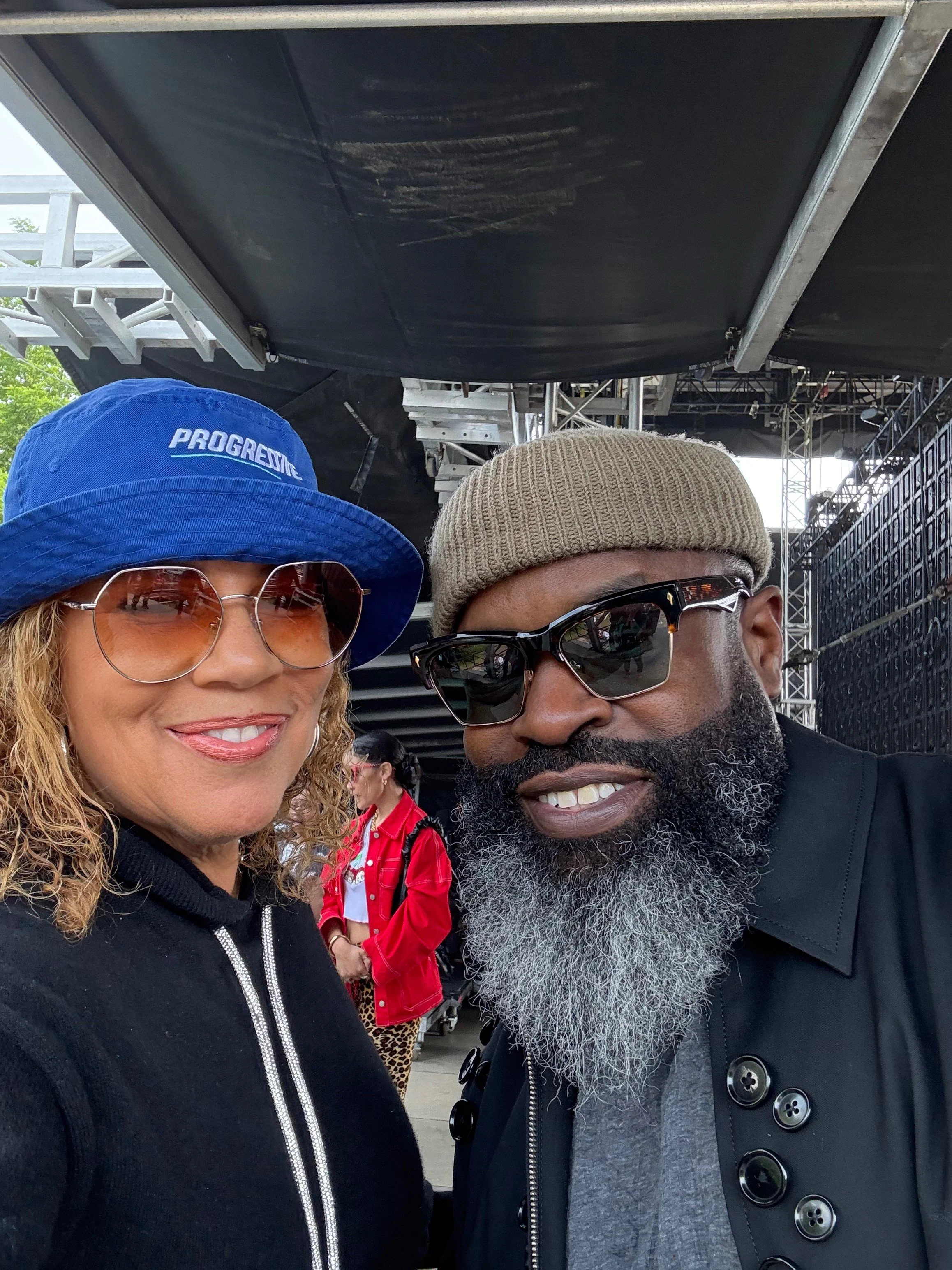 A woman with curly blonde hair wearing a blue bucket hat and sunglasses smiling next to a man with a gray beard, black sunglasses, a beige knit cap, and black clothing. They are outdoors near a stage or event setup.