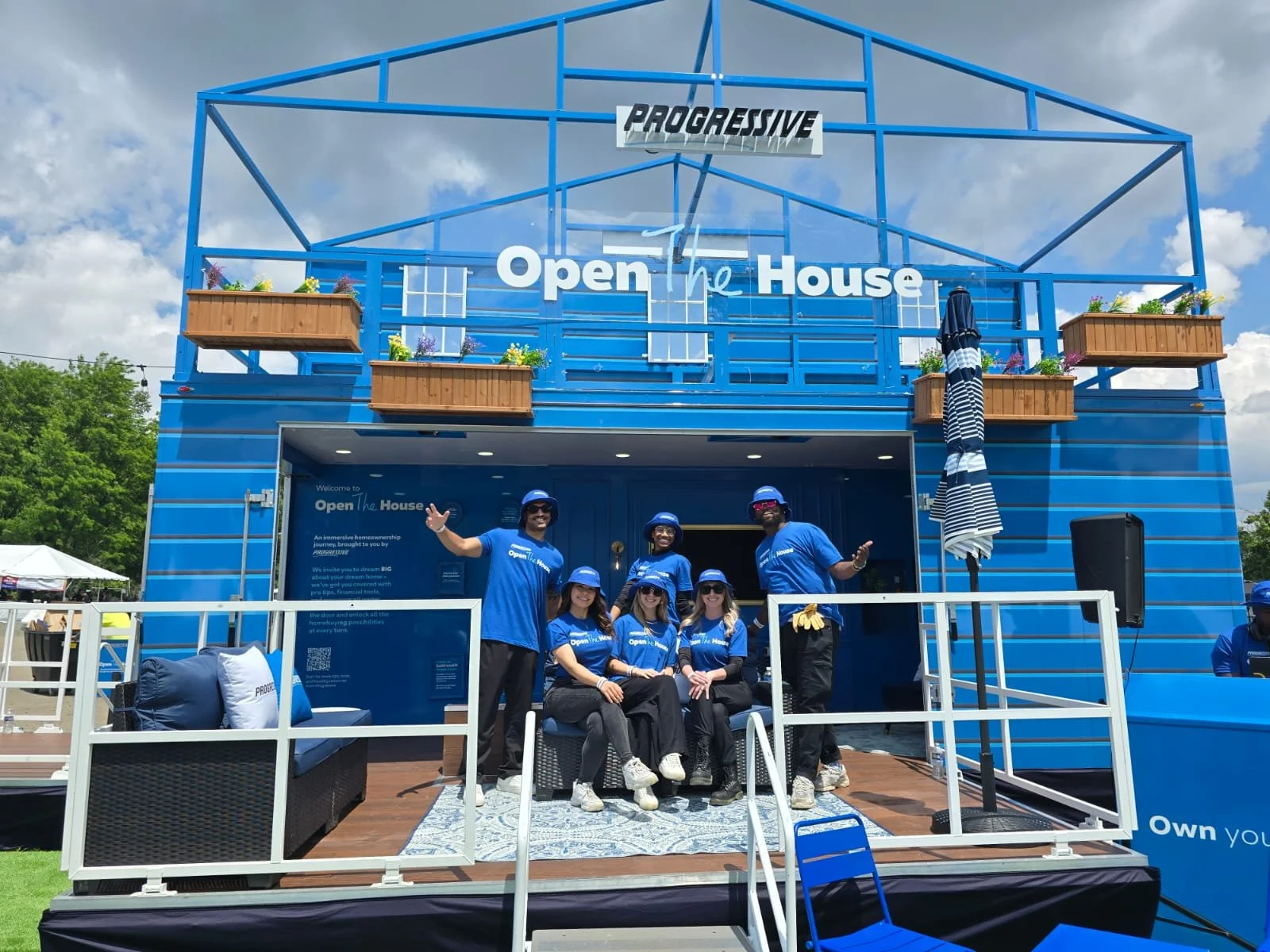 People in blue shirts and hats standing and sitting in front of a blue house with signs that say 'Open the House' and 'Progressive' at an outdoor event.