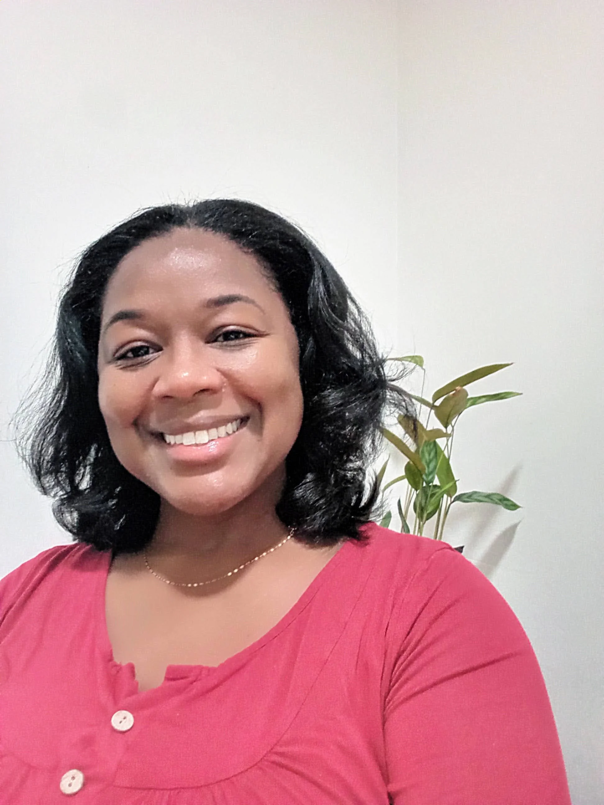 A smiling woman with shoulder-length curly black hair, wearing a pink top with buttons and a small necklace, standing in front of a white wall with a plant in the background.