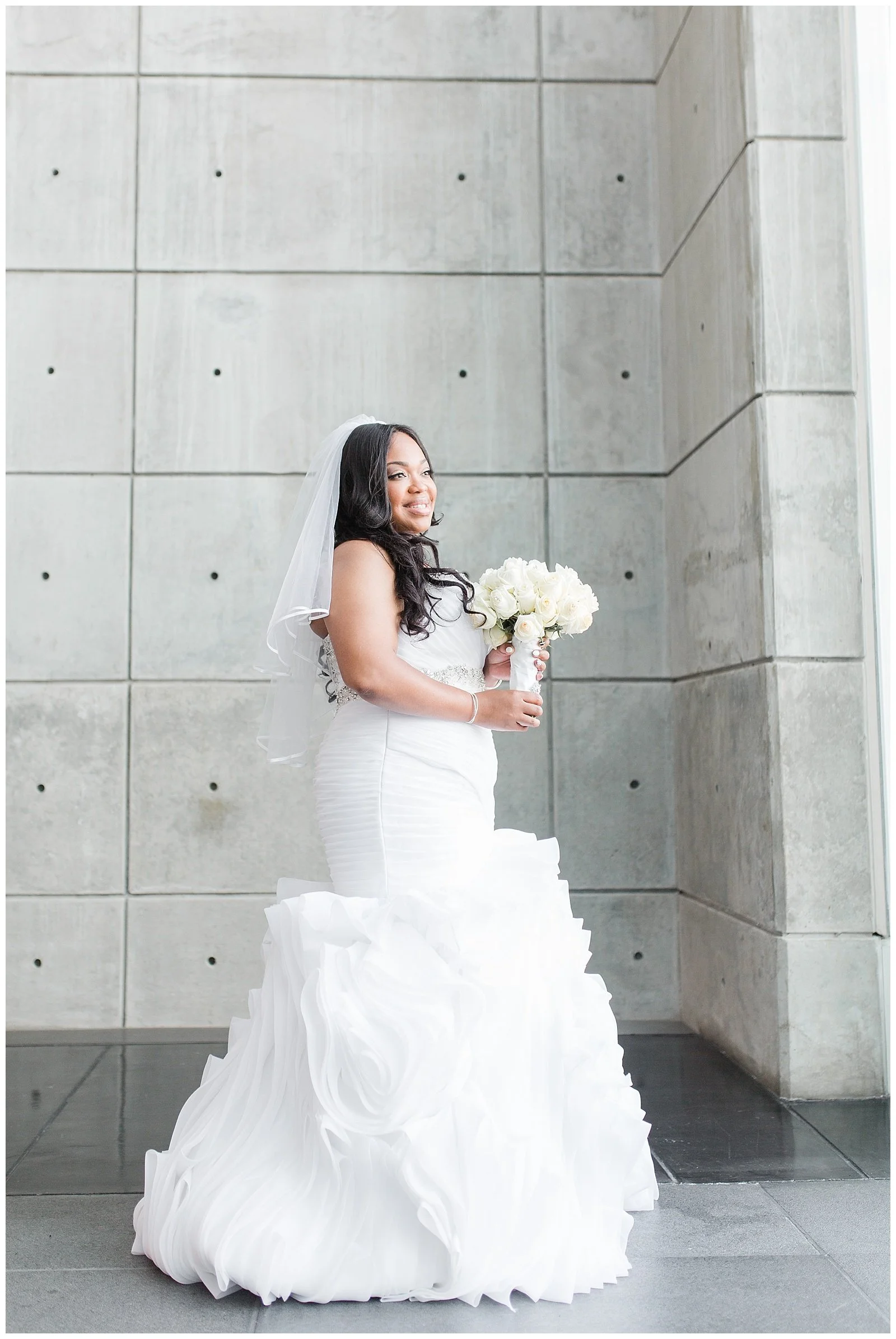A bride in a white wedding dress holding a bouquet of white roses, standing indoors against a concrete wall.