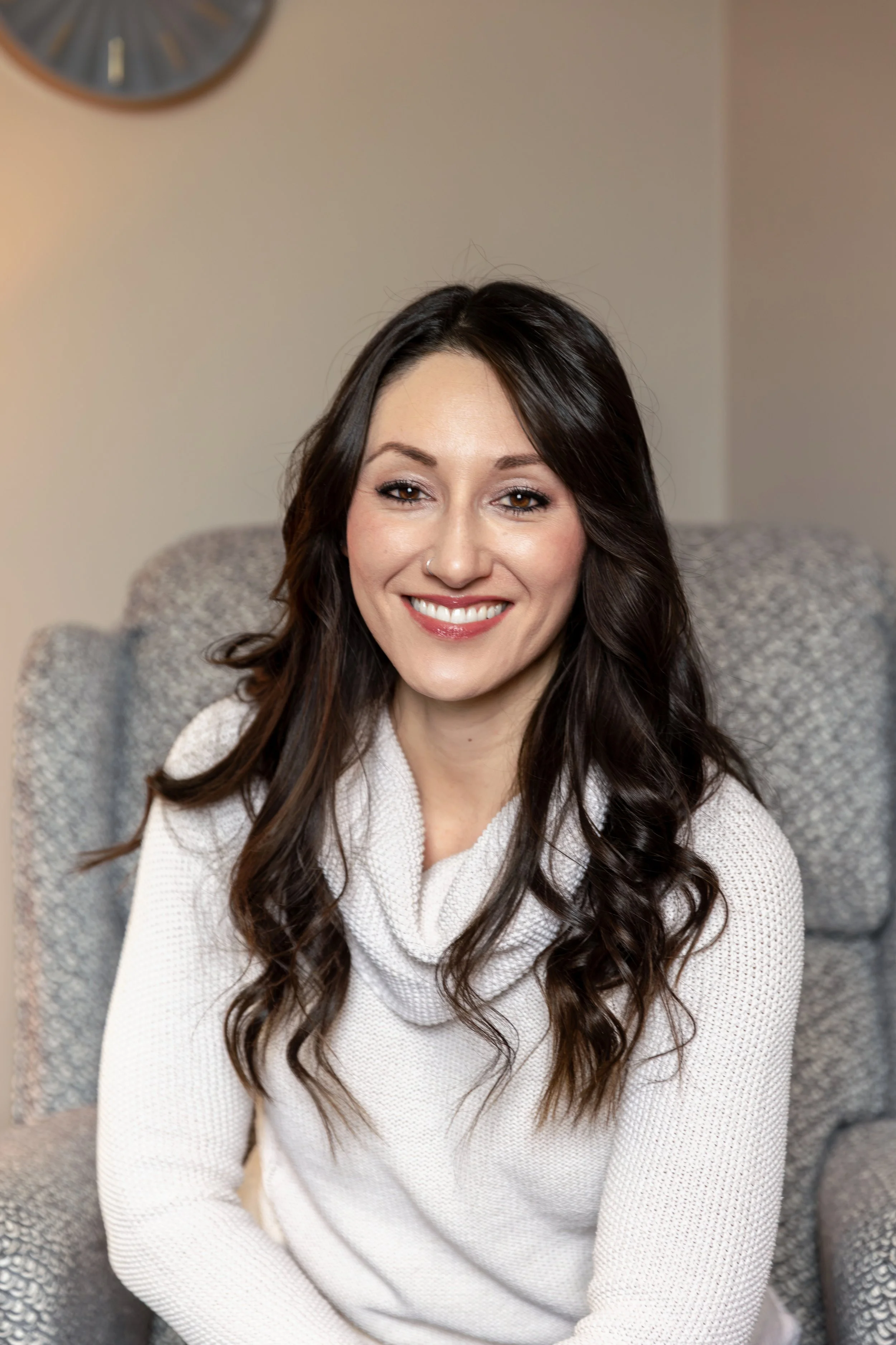A woman with long dark brown hair and a nose piercing smiling, wearing a black and white patterned sweater, standing against a plain white wall.
