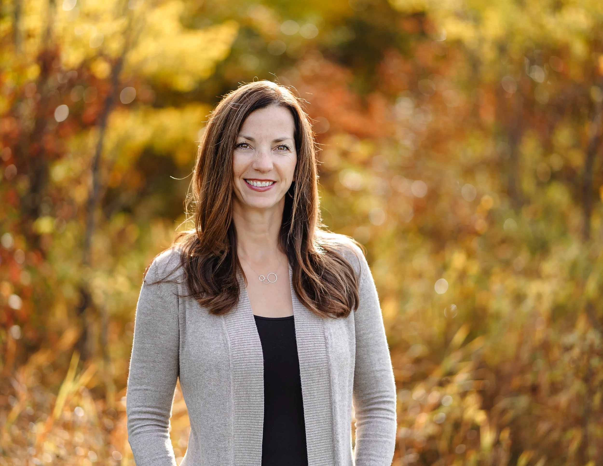 A woman with long brown hair smiling outdoors during fall with yellow and orange foliage in the background.