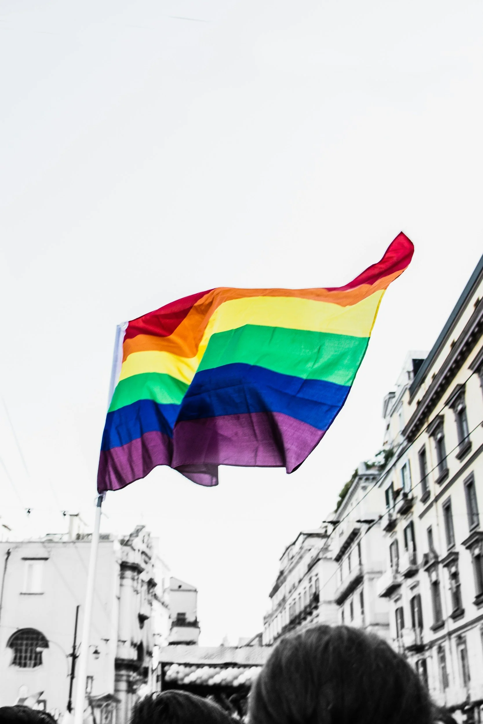A rainbow pride flag flying in the air in an urban setting with tall buildings and a crowd below.