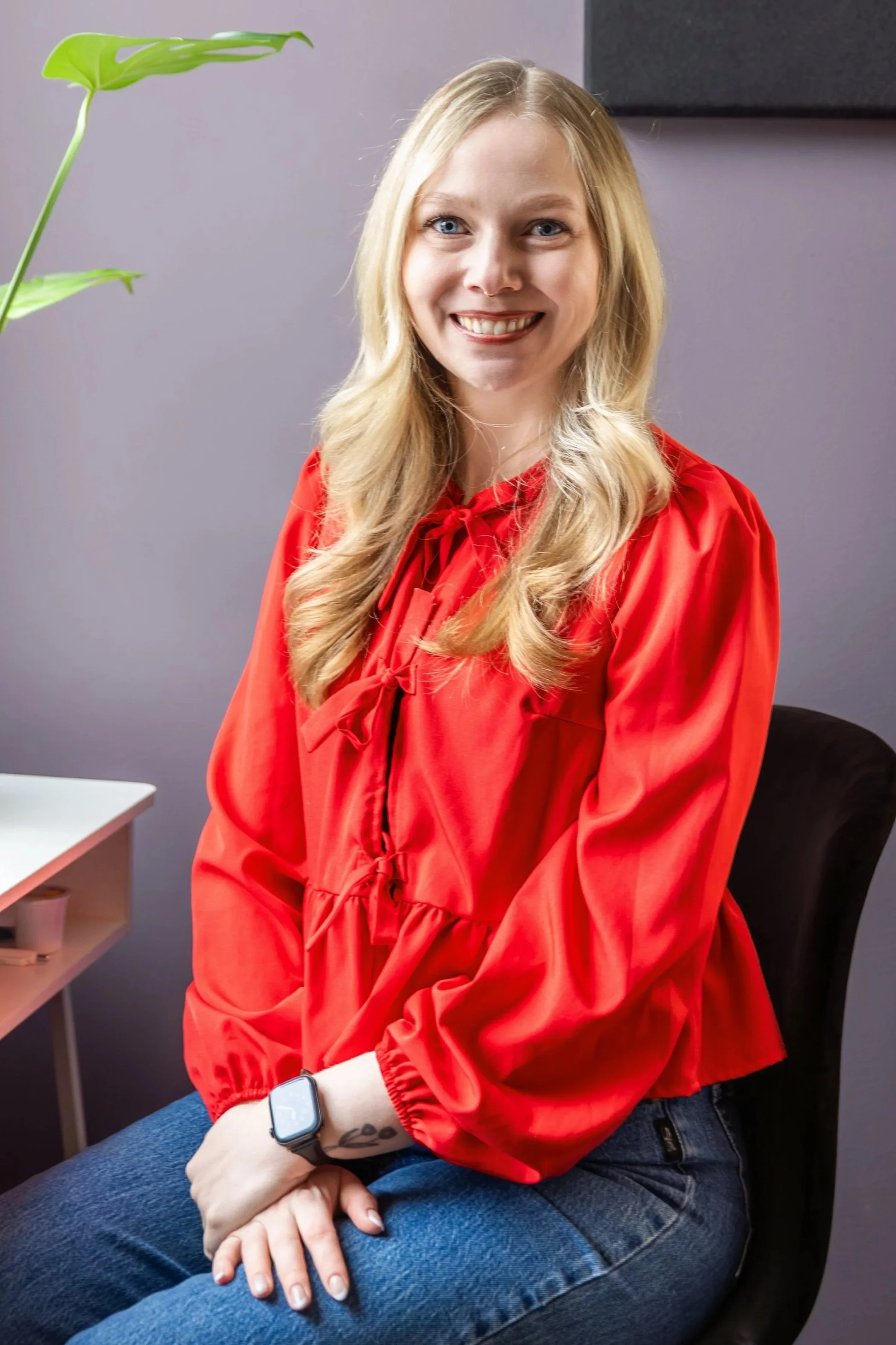 A woman with blonde hair and blue eyes wearing a red shirt, sitting on a chair, smiling, with a plant in the background and a smartwatch on her wrist.