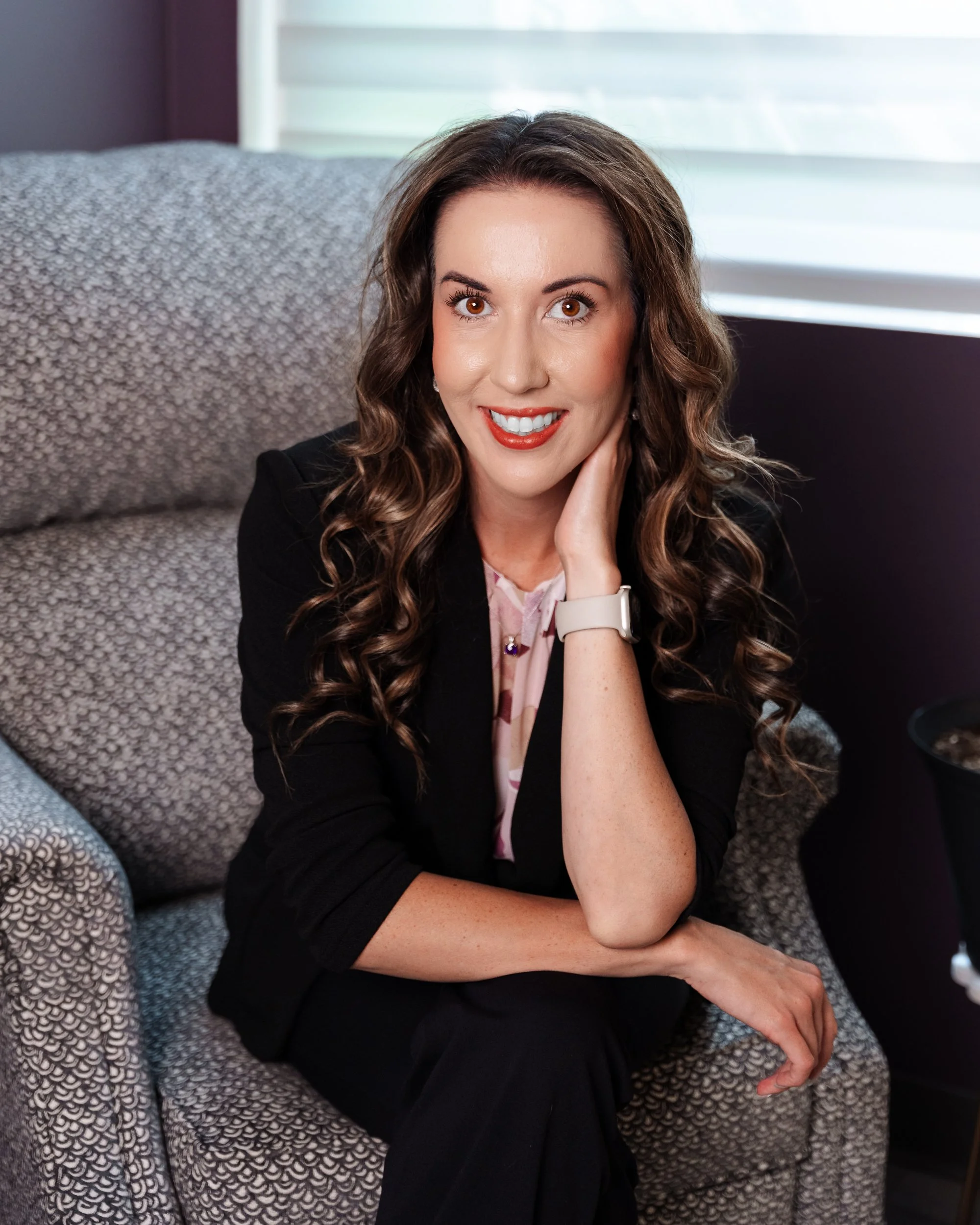A woman with long, curly brown hair, wearing a black blazer and pink blouse, smiling and resting her head on her hand while sitting on a patterned couch in a room with window blinds.