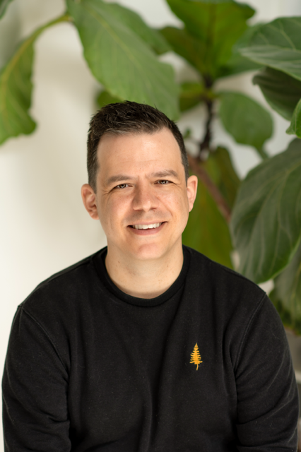 A man smiling in front of large green plant leaves, wearing a black shirt with a small yellow tree logo.