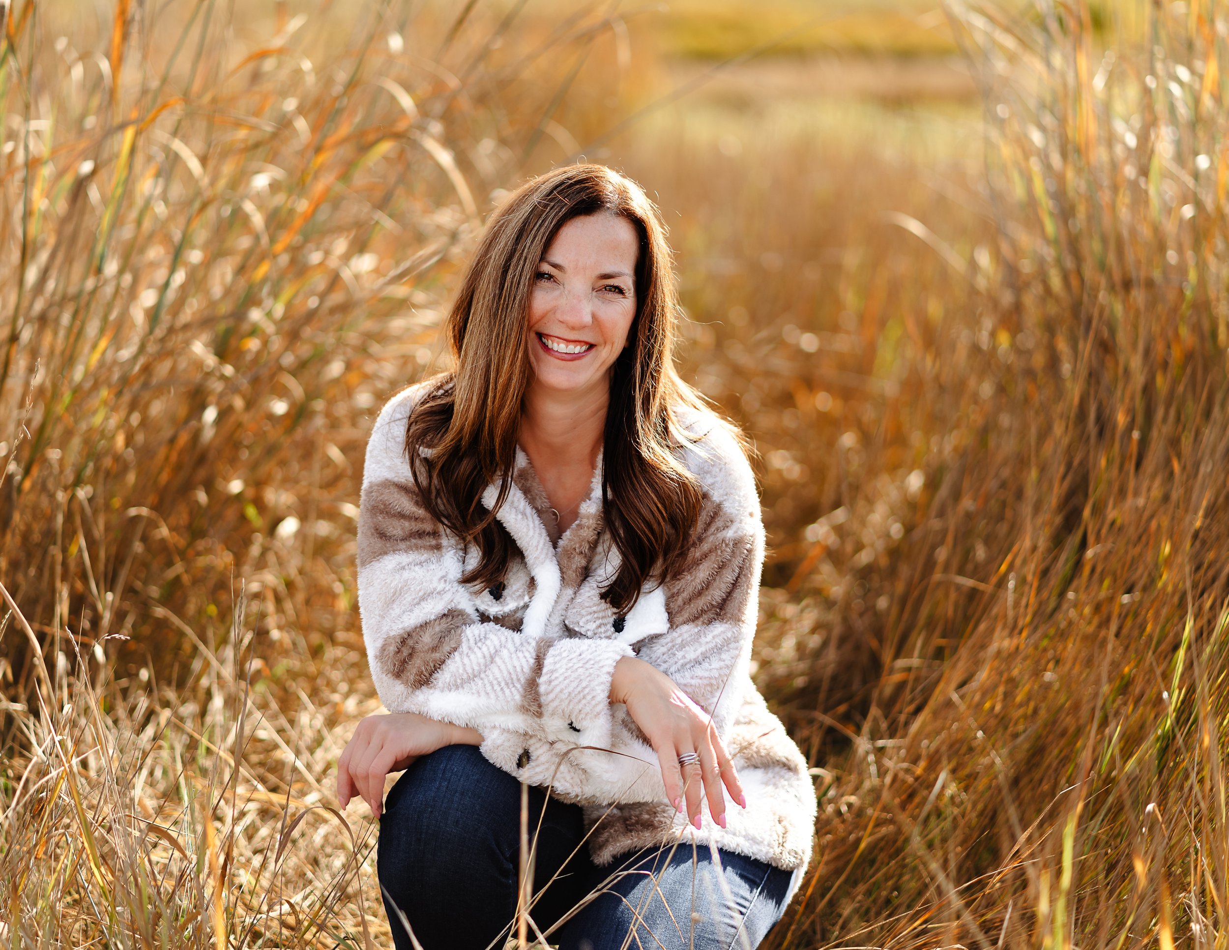 A woman with long brown hair smiling and sitting among tall, golden autumn grass.