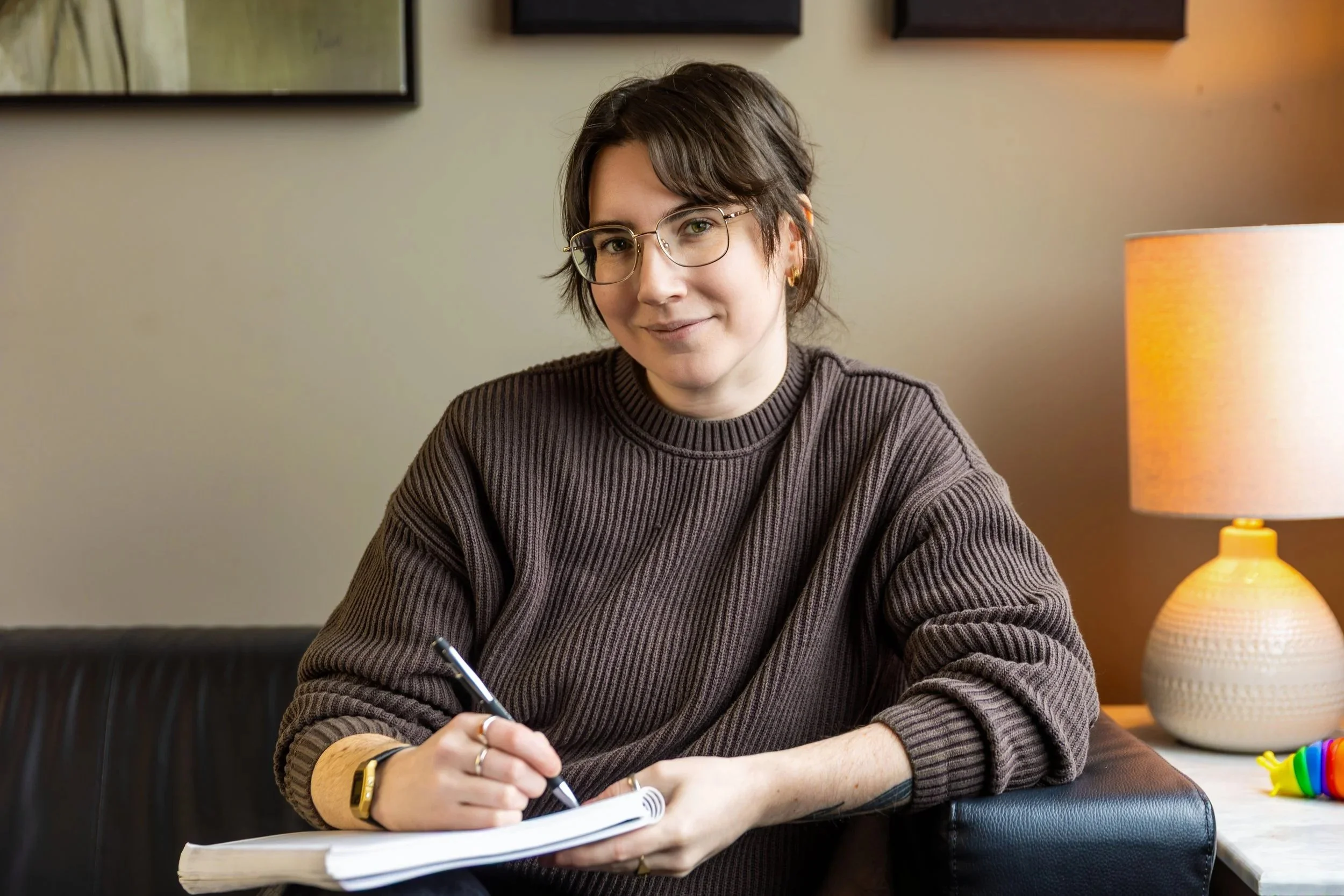 A counsellor wearing glasses sits in a warmly lit therapy office holding a pen and notebook.
