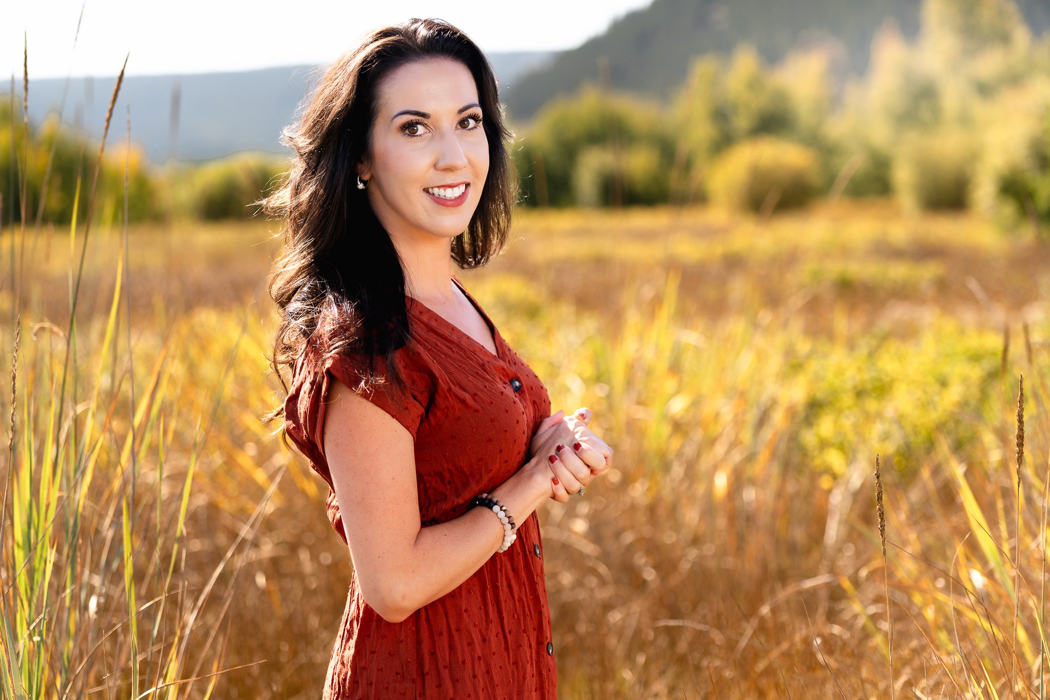 Sheri Stahl with dark hair and fair skin standing in a field of tall, yellow grass during daytime, wearing a red dress and smiling at the camera.