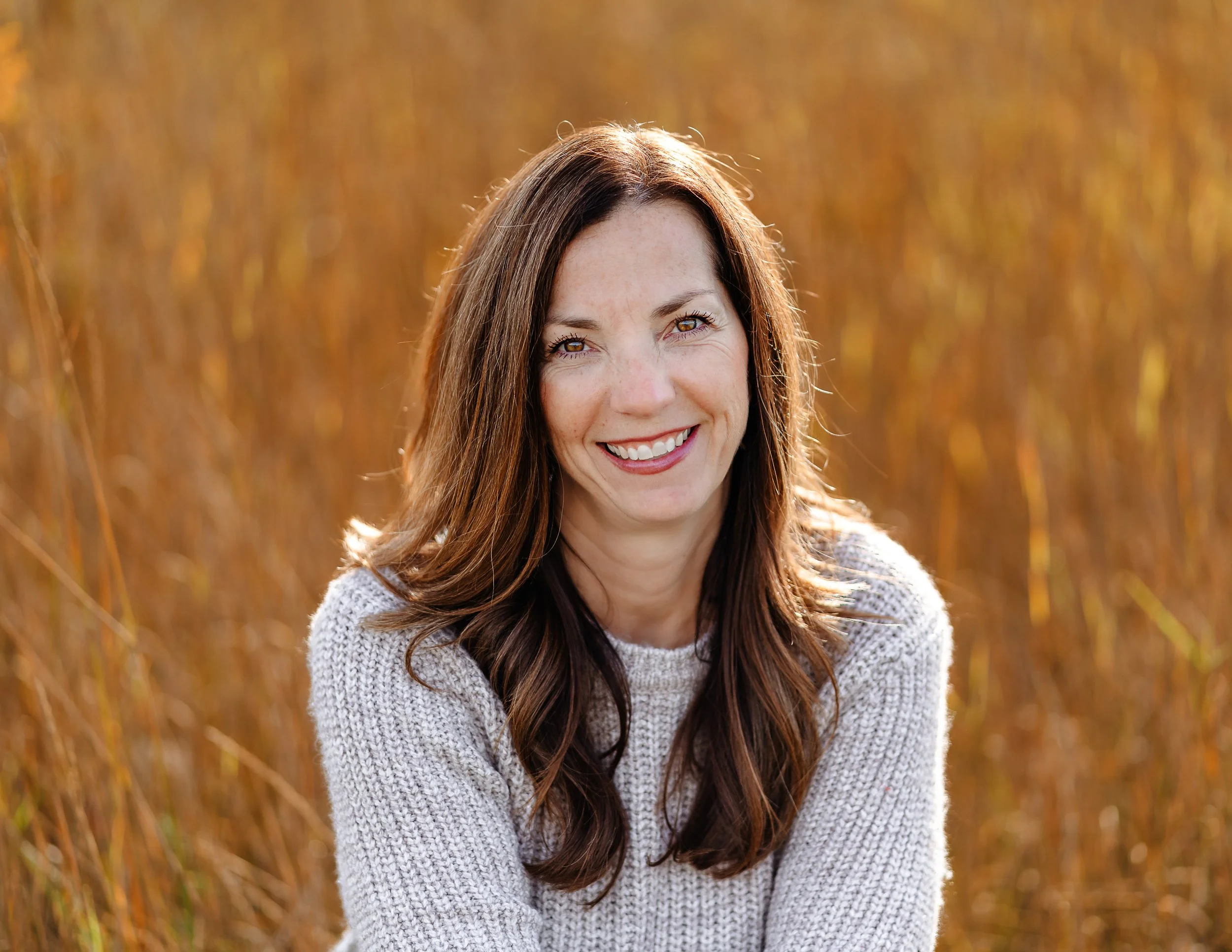 A woman with long brown hair smiling and resting her hand on her cheek, sitting on a grey couch with pink pillows, wearing a floral top with cutout shoulders.