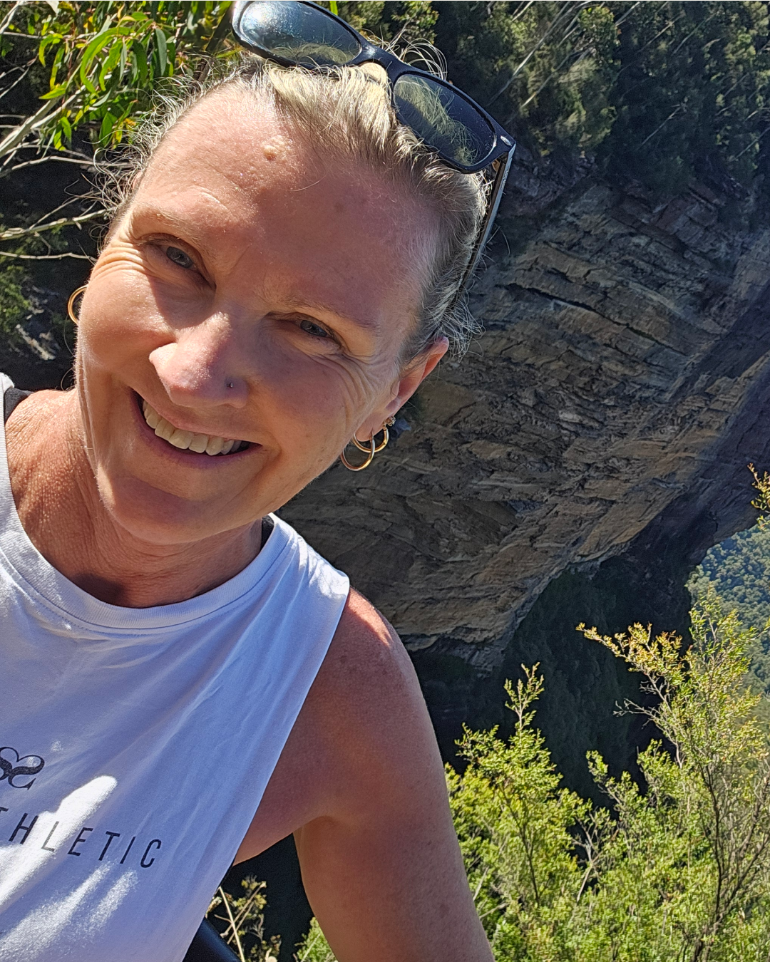 A woman smiling outdoors with sunglasses on her head, wearing a white athletic tank top, and standing in front of a large cliff with green vegetation around. Yoga in the Blue Mountains.