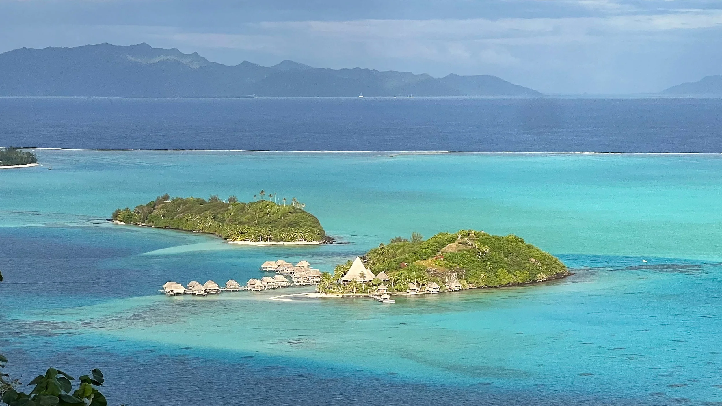 Aerial view of Bora Bora hotel with overwater bungalows in turquoise lagoon. Raiatea visible beyond the reef.